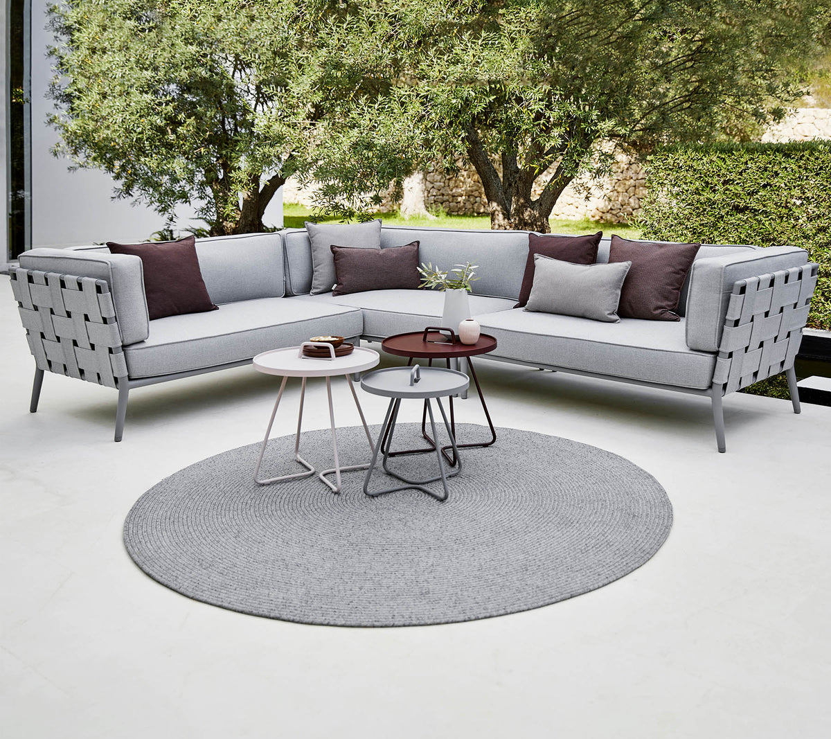 Canapé sectionnel moderne en gris clair avec des coussins décoratifs, accompagné de tables basses élégantes sur un tapis rond. Verdure luxuriante en arrière-plan.