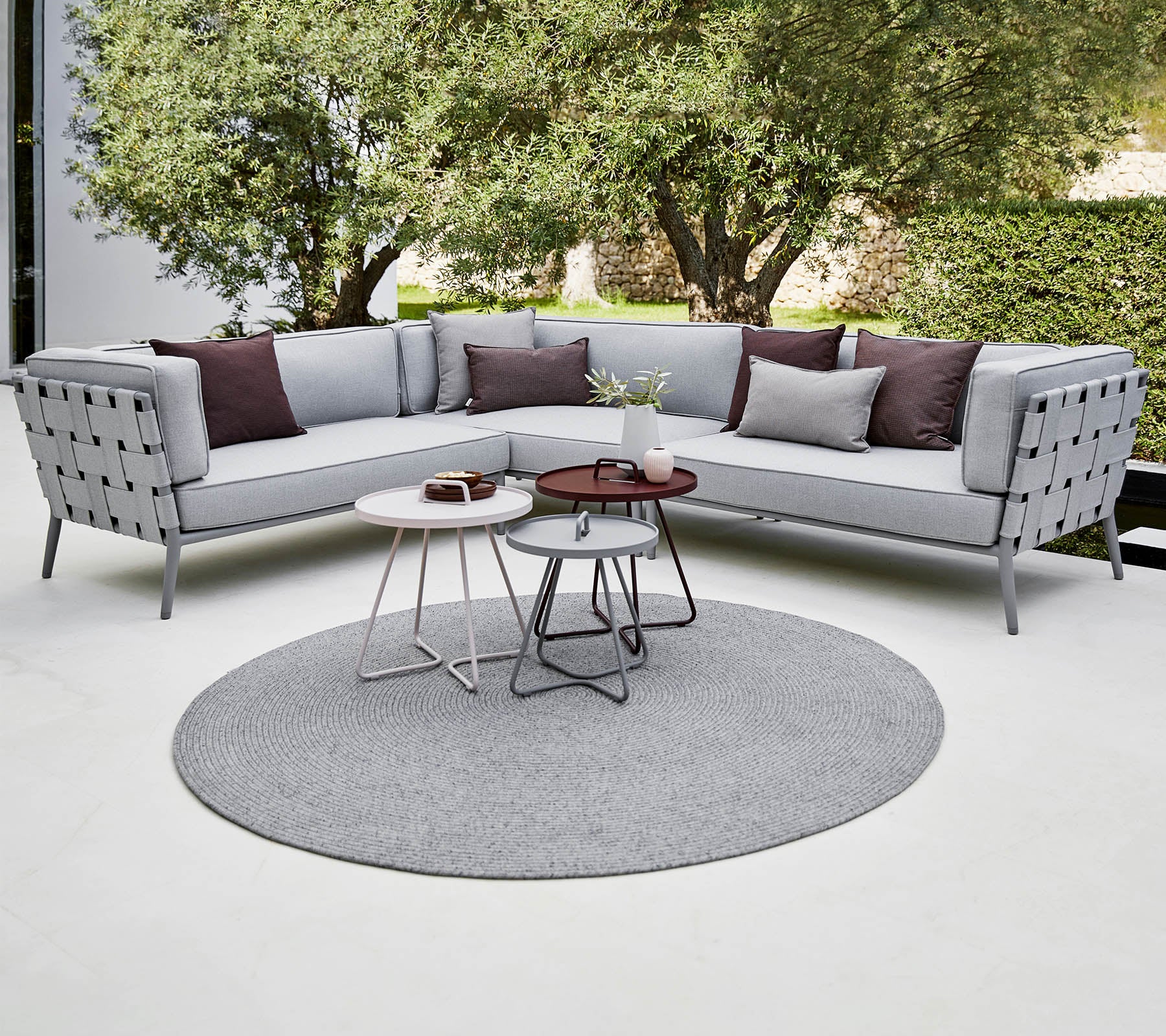 Canapé sectionnel moderne en gris clair avec des coussins décoratifs, accompagné de tables basses élégantes sur un tapis rond. Verdure luxuriante en arrière-plan.