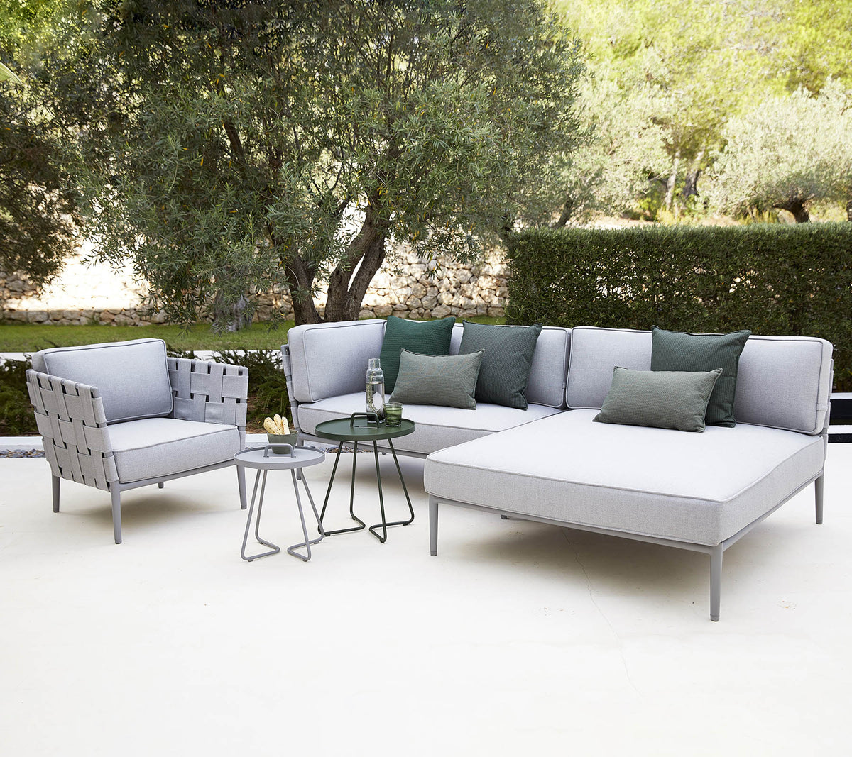 Agencement moderne de sièges extérieurs avec un canapé sectionnel avec des coussins, une chaise et une petite table en verre entourée de verdure.