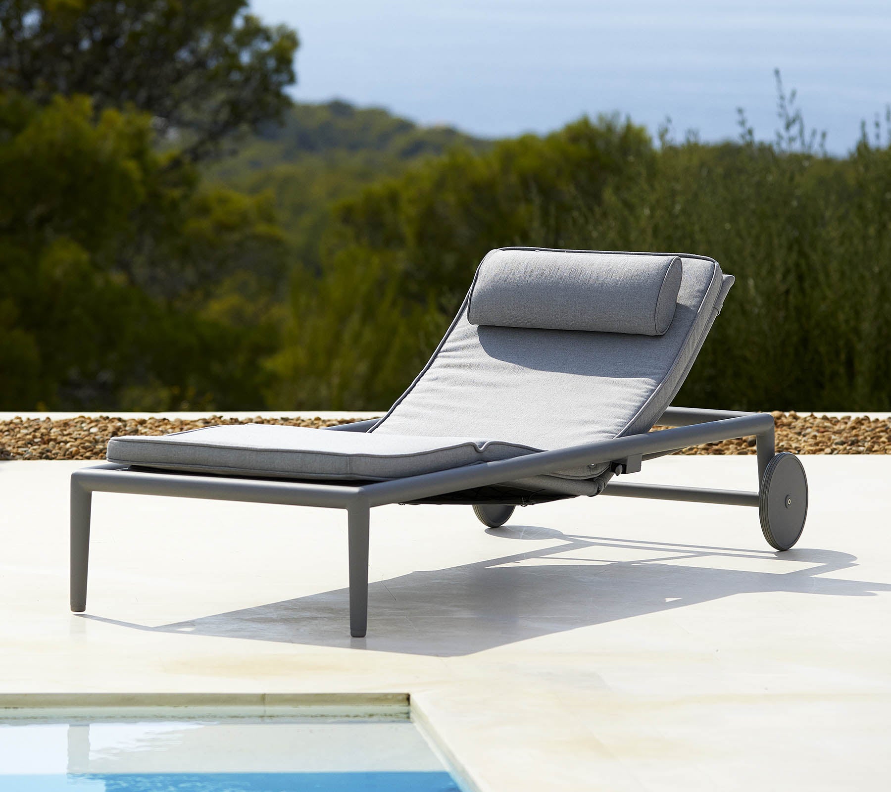 Fauteuil lounge confortable avec un coussin gris et un dossier, équipé de roues pour une mobilité facile, à côté d'une piscine et de verdure luxuriante en arrière-plan.