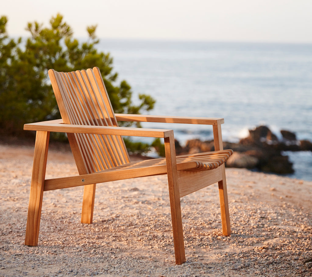 Vue de côté d'une chaise longue en bois avec un dossier à lattes, positionnée près d'une scène aquatique sereine et entourée de verdure.