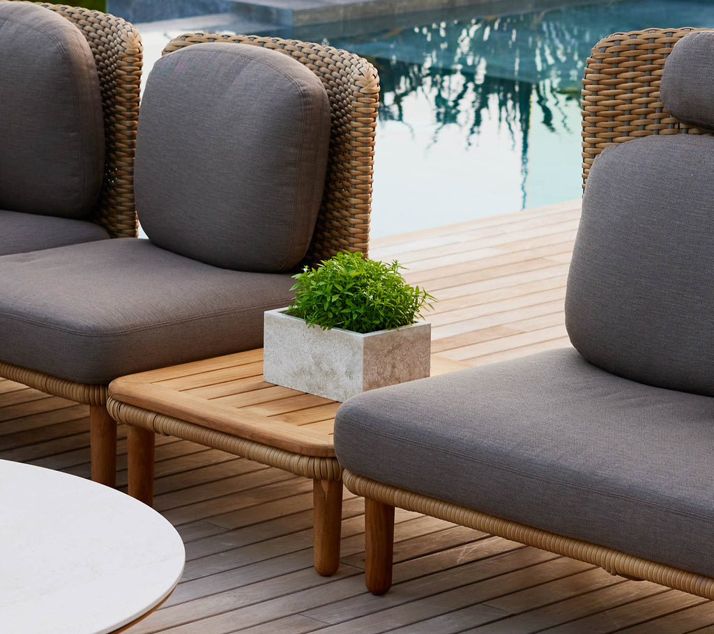Une table basse Arch élégante avec un plateau en teck, flanquée de chaises longues et d'une petite plante en pot, améliorant un cadre extérieur chic.