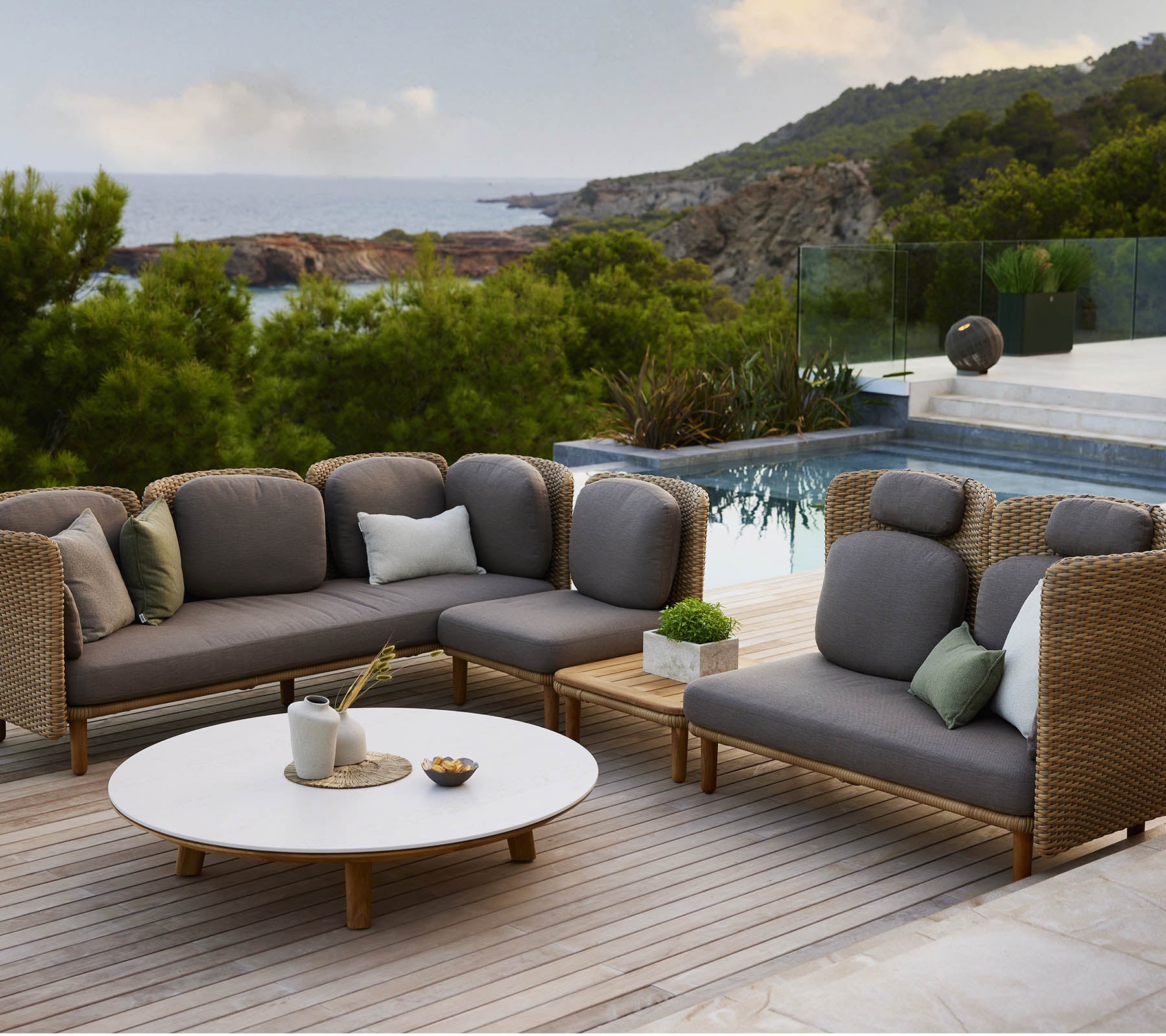 Mobilier de salon élégant avec des coussins gris et des coussins décoratifs, accompagné d'une table ronde, sur fond de paysage pittoresque.