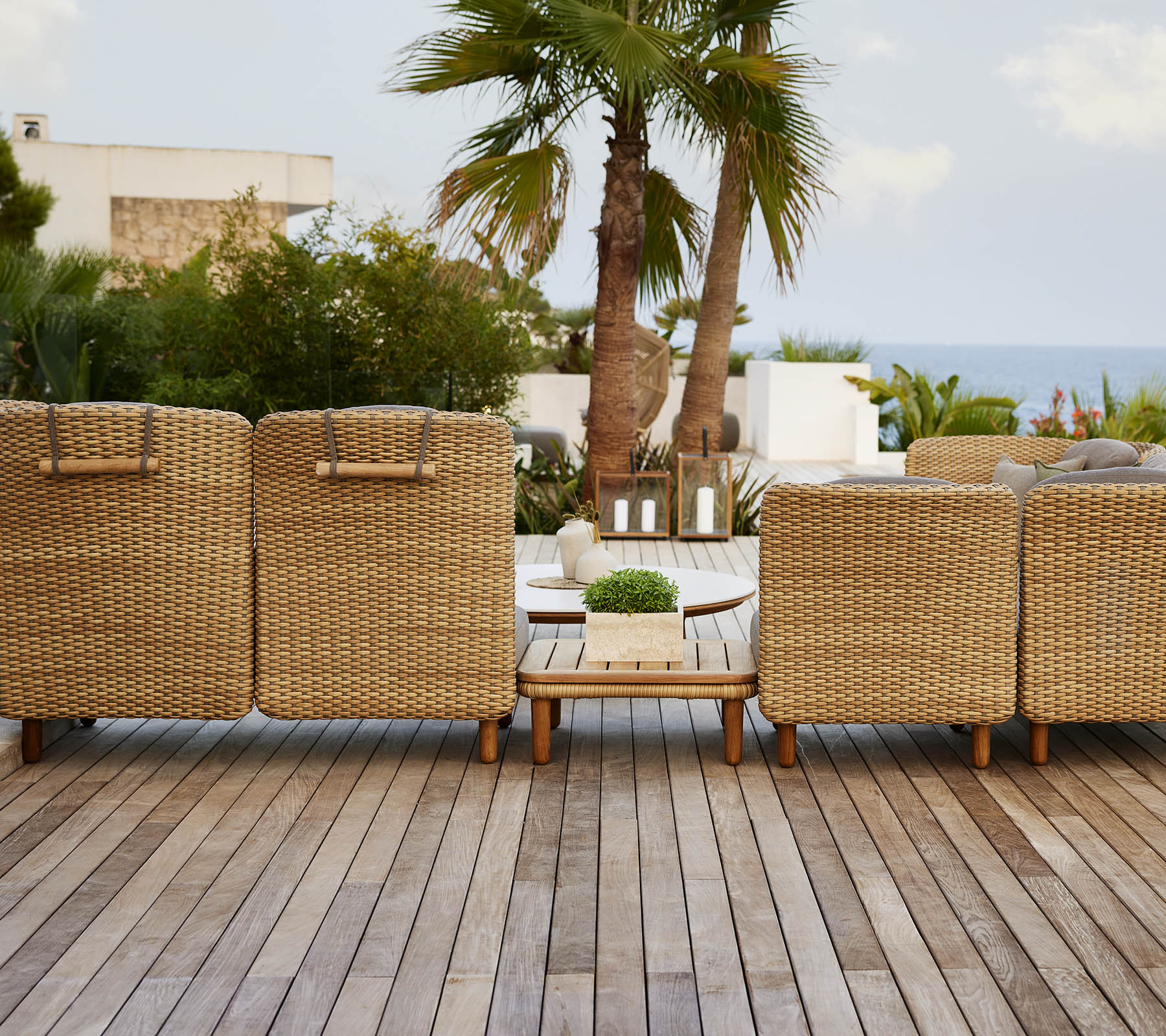 Une rangée de chaises longues tissées faisant face à une vue pittoresque, avec une terrasse en bois et des plantes tropicales en arrière-plan.