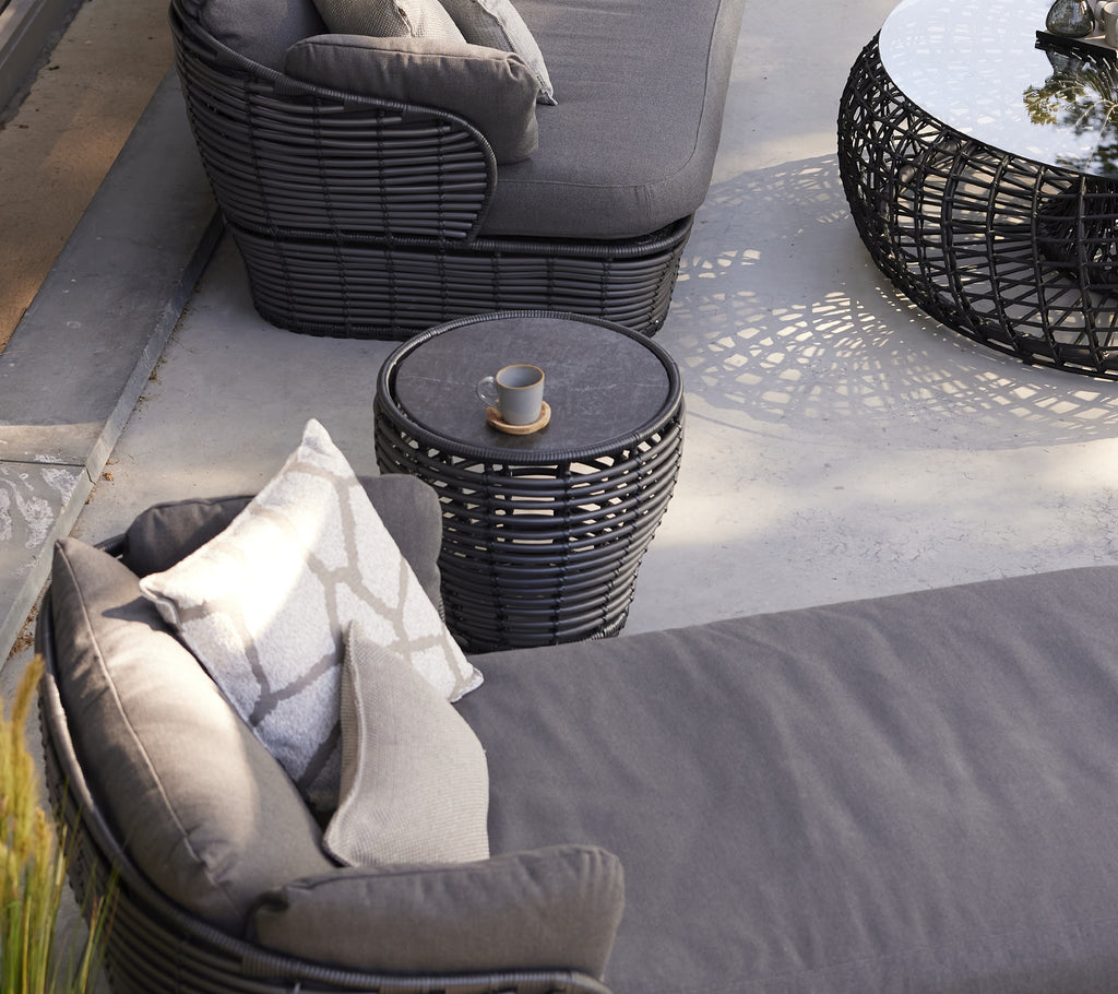Une petite table basse en graphite en forme de panier à côté d'une chaise longue moderne avec des coussins, améliorant un espace de vie élégant.