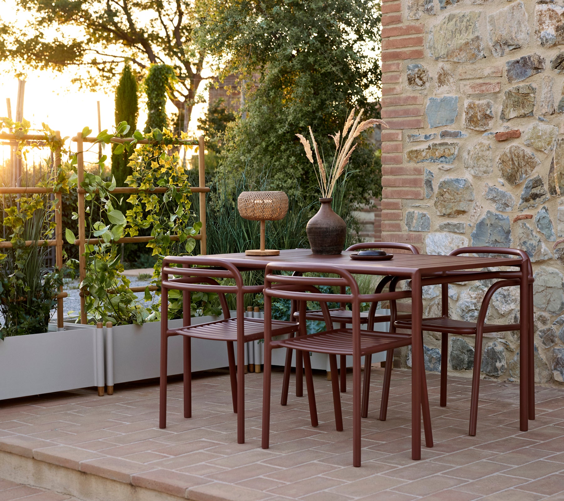 Cadre extérieur avec une table Bliss et des chaises, un fauteuil Bliss et un pot de fleurs Sticks entouré de verdure au coucher du soleil.