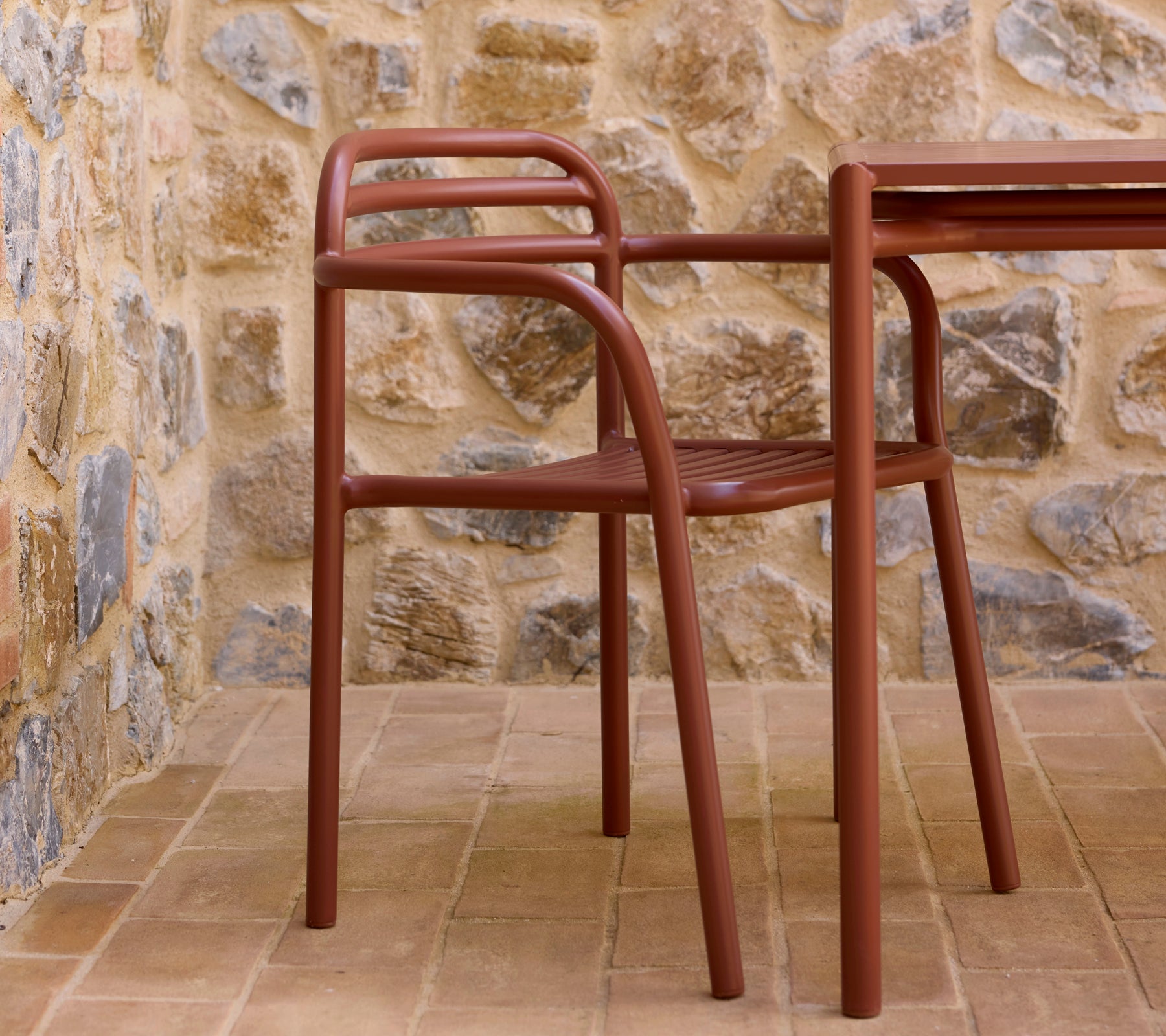 Fauteuil Bliss rouge désert à côté d'une table assortie, placé contre un mur en pierres texturées. Un agencement d'assise à la fois élégant et moderne.