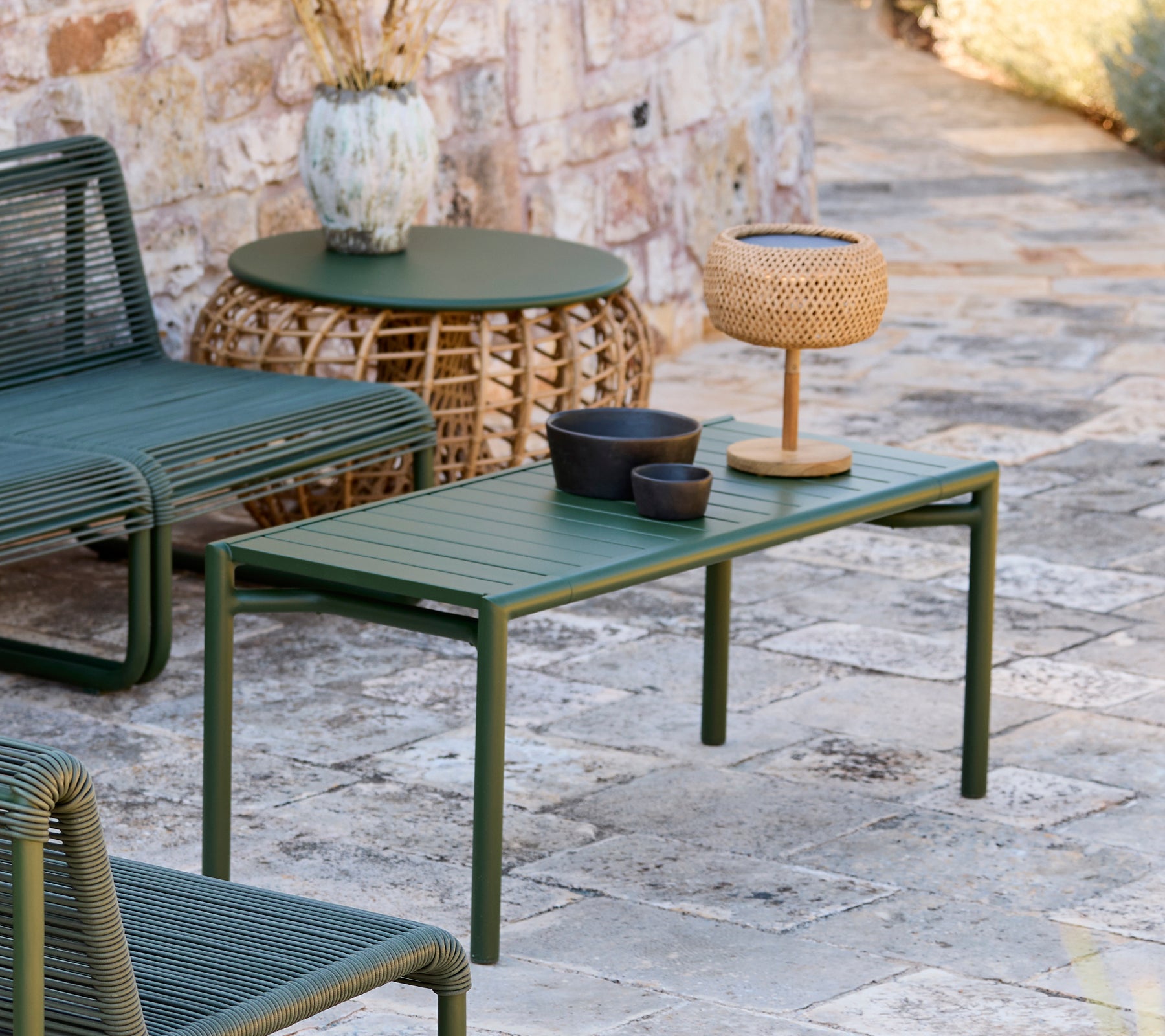 Table verte avec bol noir, vase marron et lampe tissée dans un espace lounge moderne en extérieur.