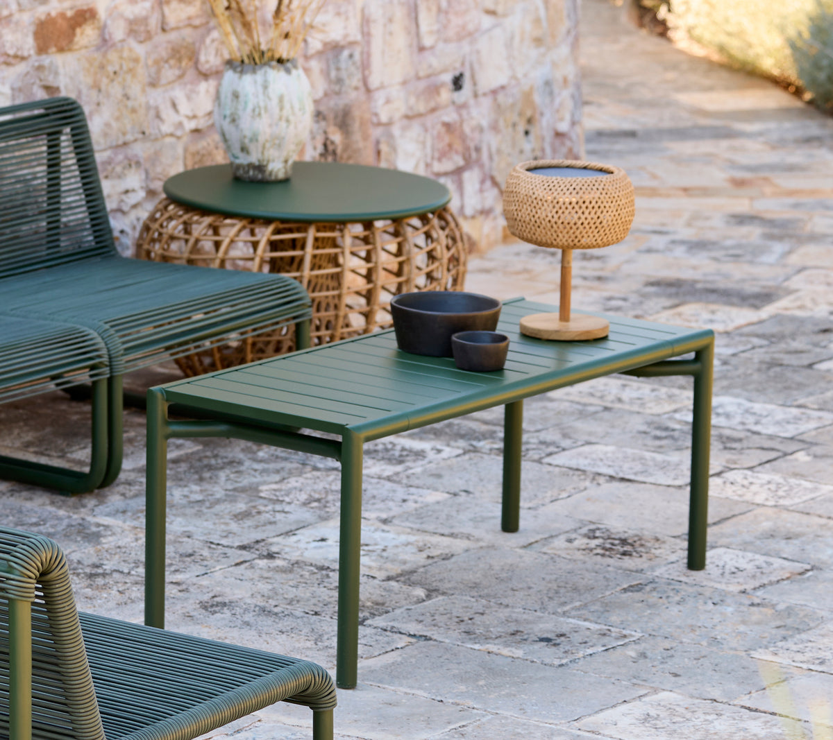 Table verte avec bol noir, vase marron et lampe tissée dans un espace lounge moderne en extérieur.