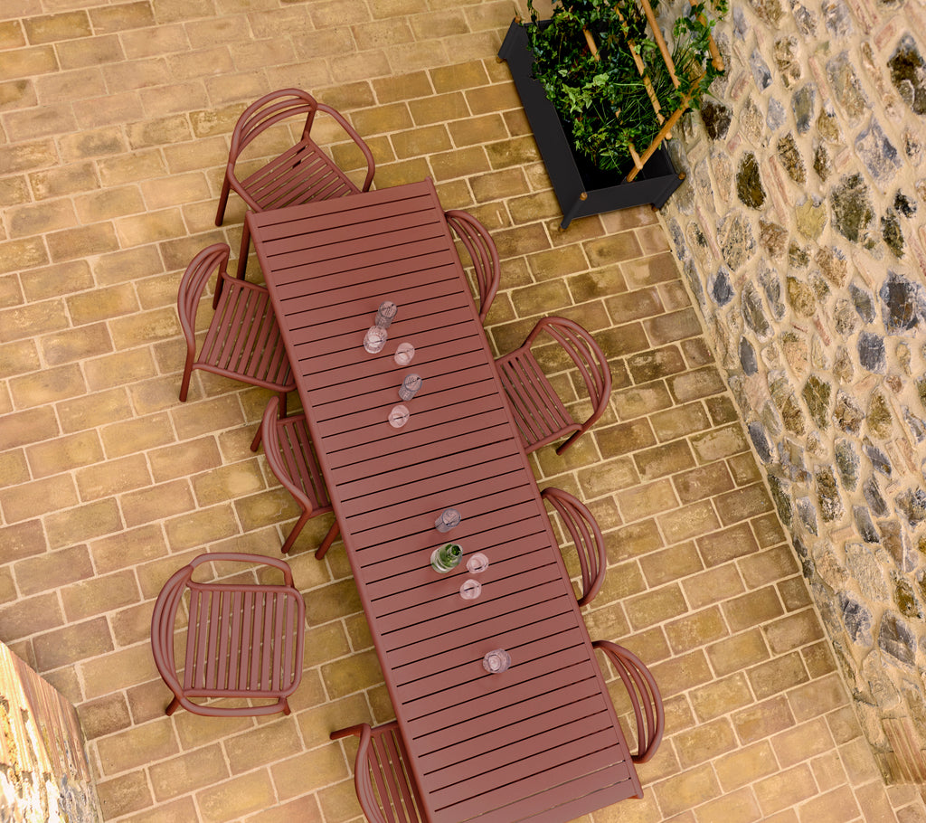 Une table Bliss rouge désert avec des chaises assorties disposées autour, placée sur un sol en pierre avec un pot de fleurs en arrière-plan.