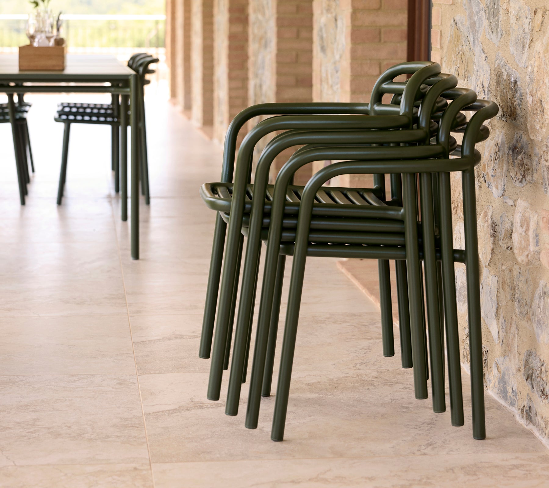 Chaises Bliss empilées dans un cadre extérieur, avec un design épuré et une couleur verte terreuse contre un mur en pierre.