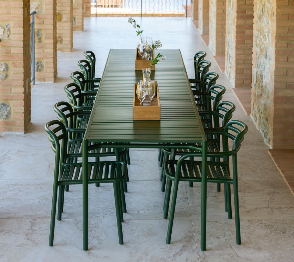 Chaises Bliss avec accoudoirs en vert foncé disposées autour d'une longue table extensible présentant un plateau décoratif central avec des fleurs.