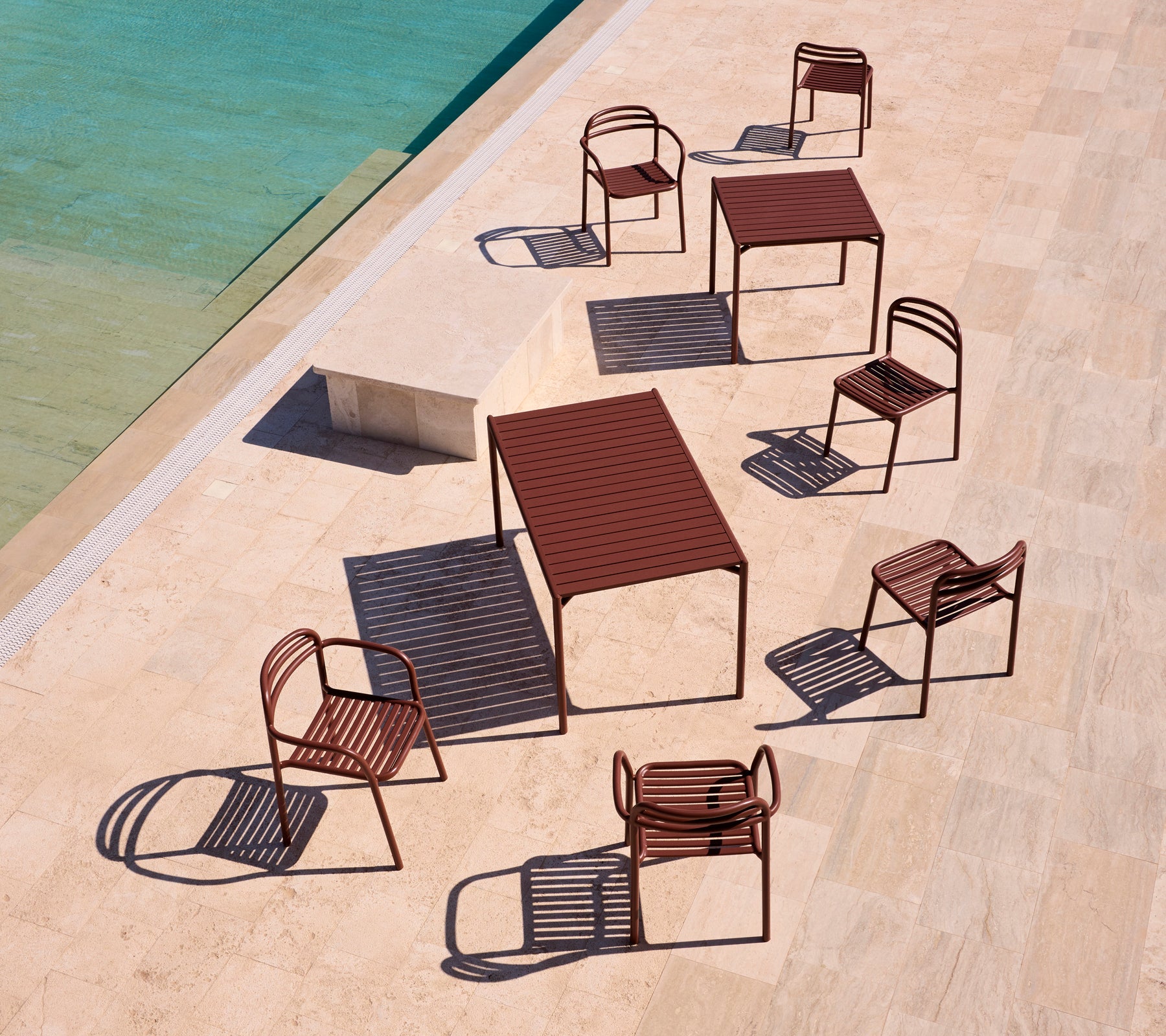 Mobilier d'extérieur de la série Bliss en rouge désert, disposé autour d'une piscine sur une terrasse ensoleillée, projetant des ombres ludiques.