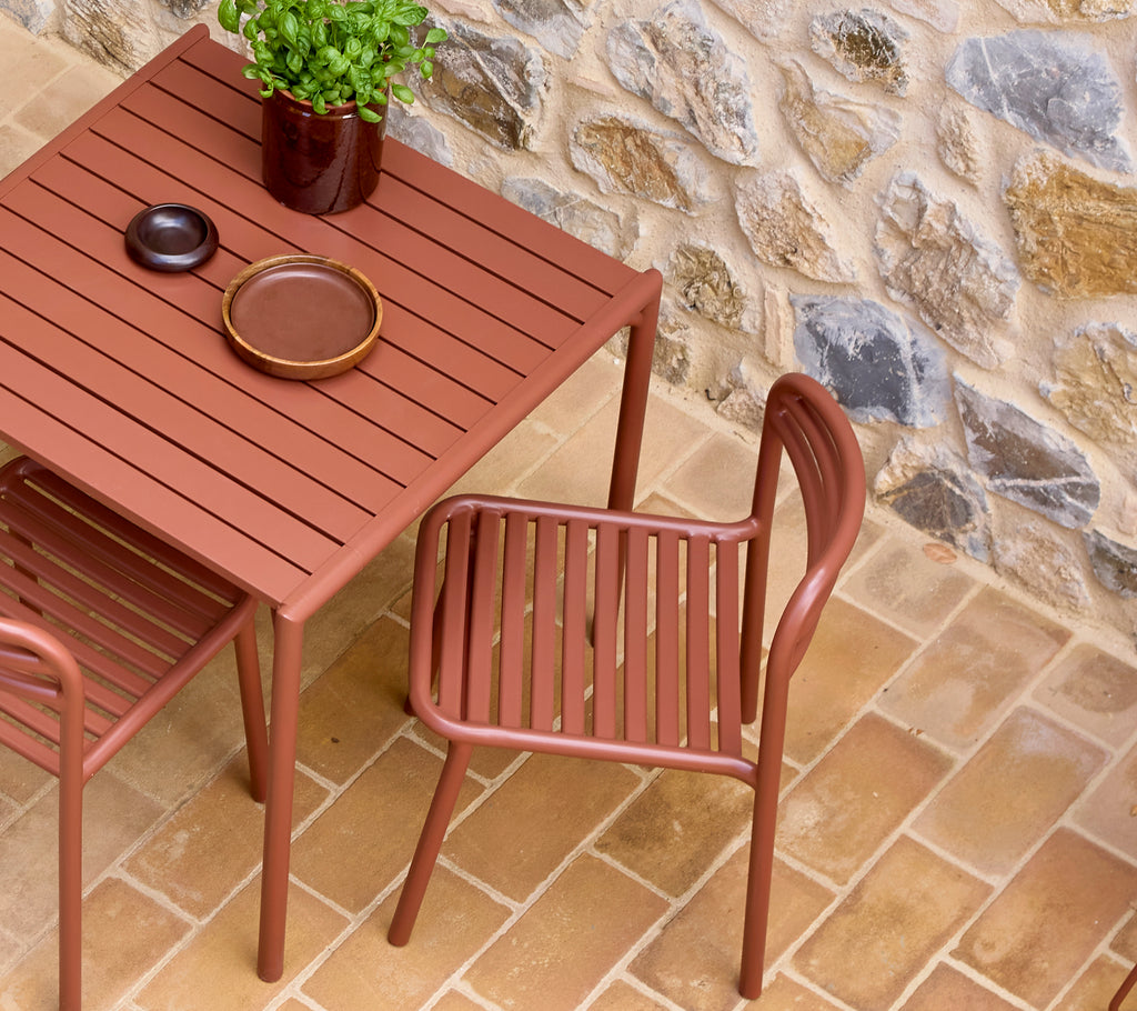 A brown metal cafe table with slatted surface, accompanied by a matching chair, small plate, and potted plant on a stone floor.