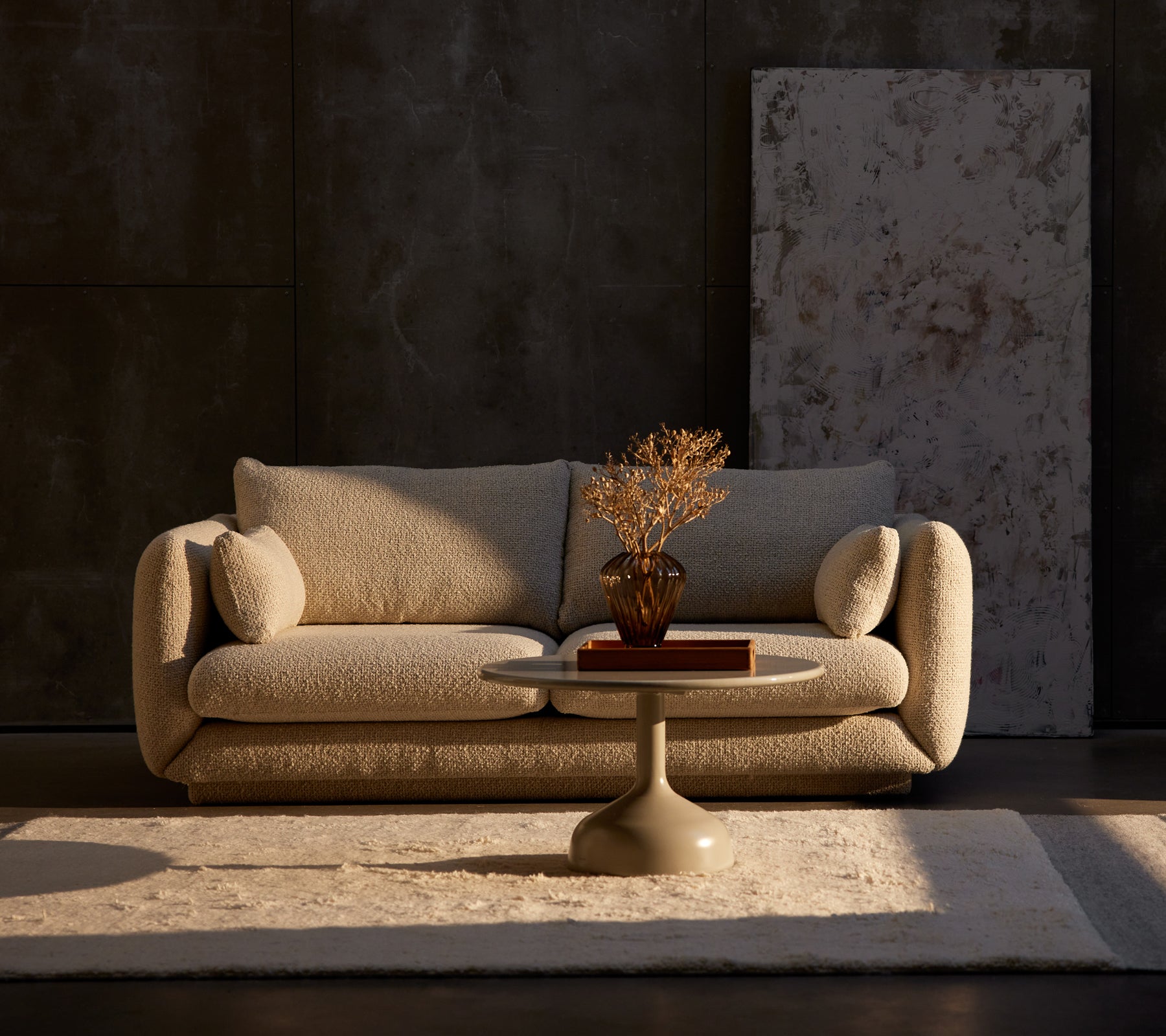 Canapé Bloom 2 places en sable flair avec une table basse Glaze et une plante décorative sur un tapis, contre un mur sombre texturé.