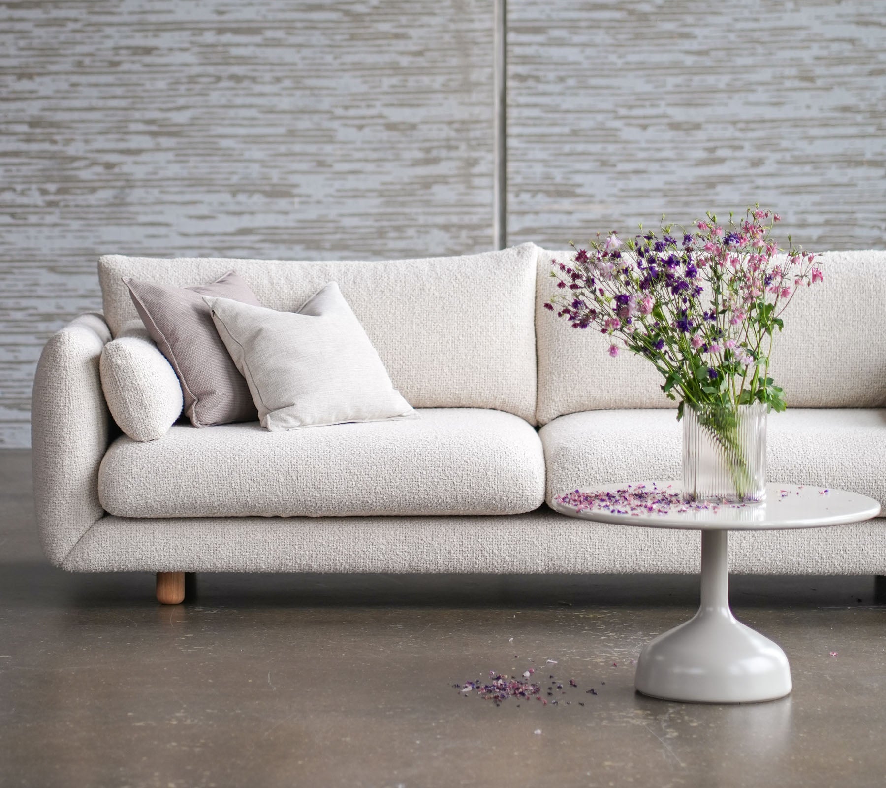 Canapé clair avec des pieds en teck, orné de coussins, complété par une table ronde et un vase de fleurs fraîches.