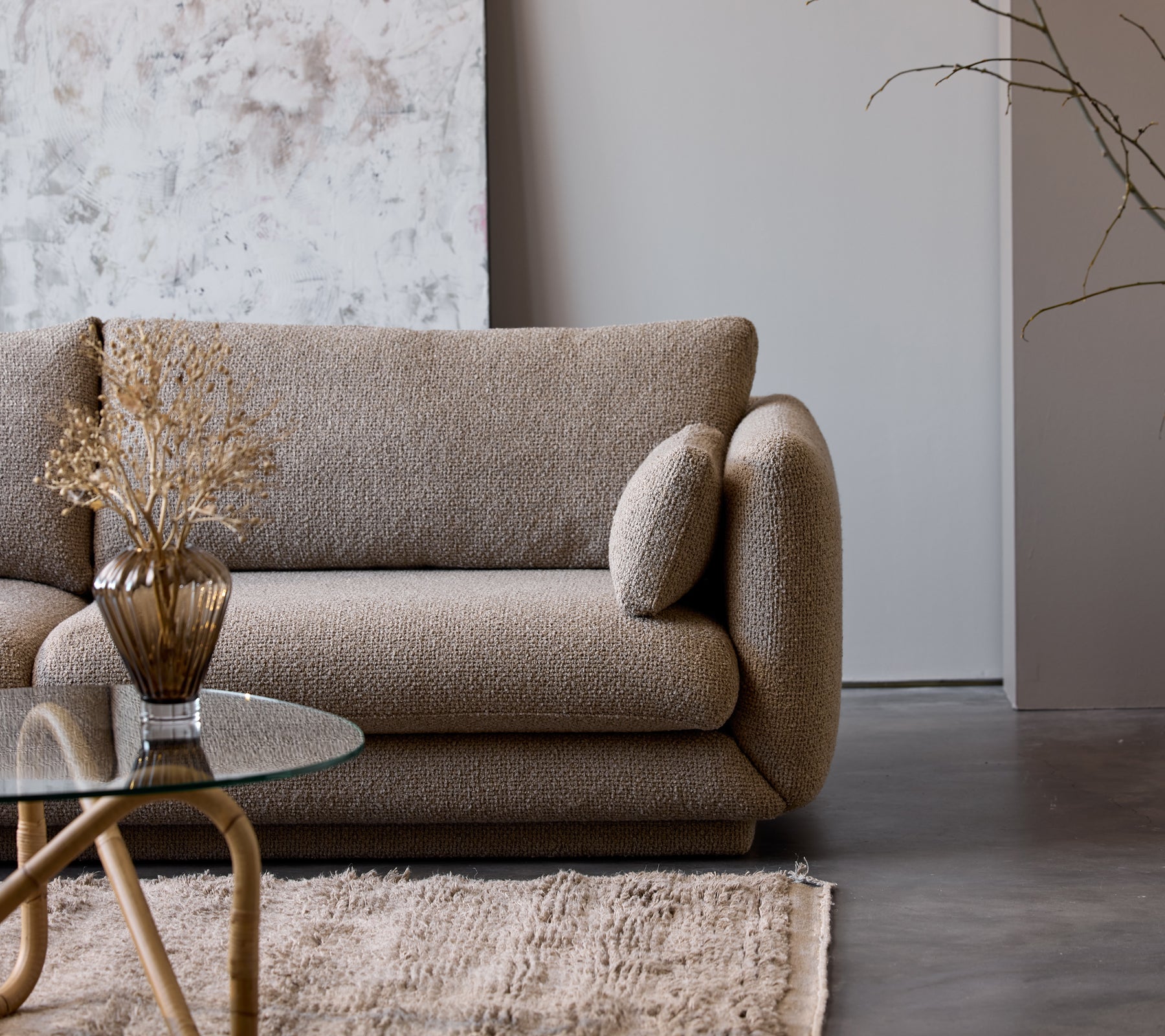 Une table basse taupe avec un plateau en verre se trouve à côté d'un canapé beige texturé orné d'un vase décoratif et de fleurs séchées.