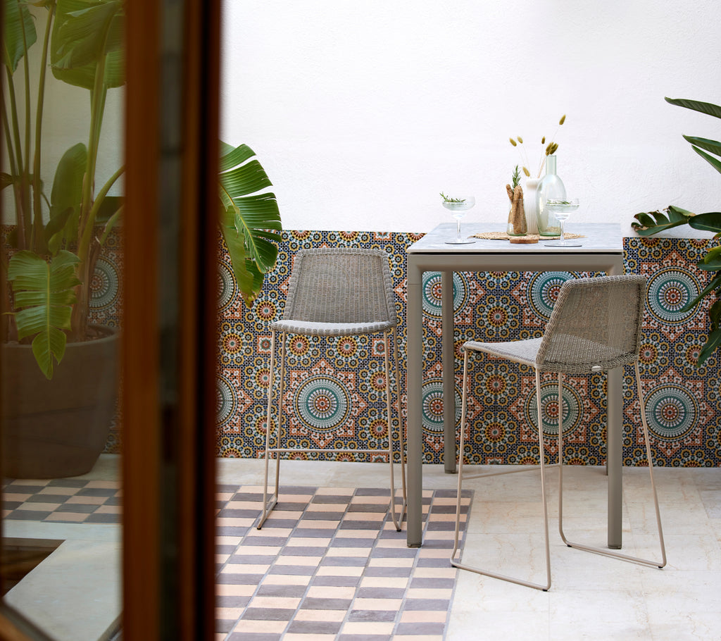 Chaises de bar Breeze en taupe avec une table de bar élégante en forme de goutte, entourées de murs décoratifs en carrelage et de plantes luxuriantes.