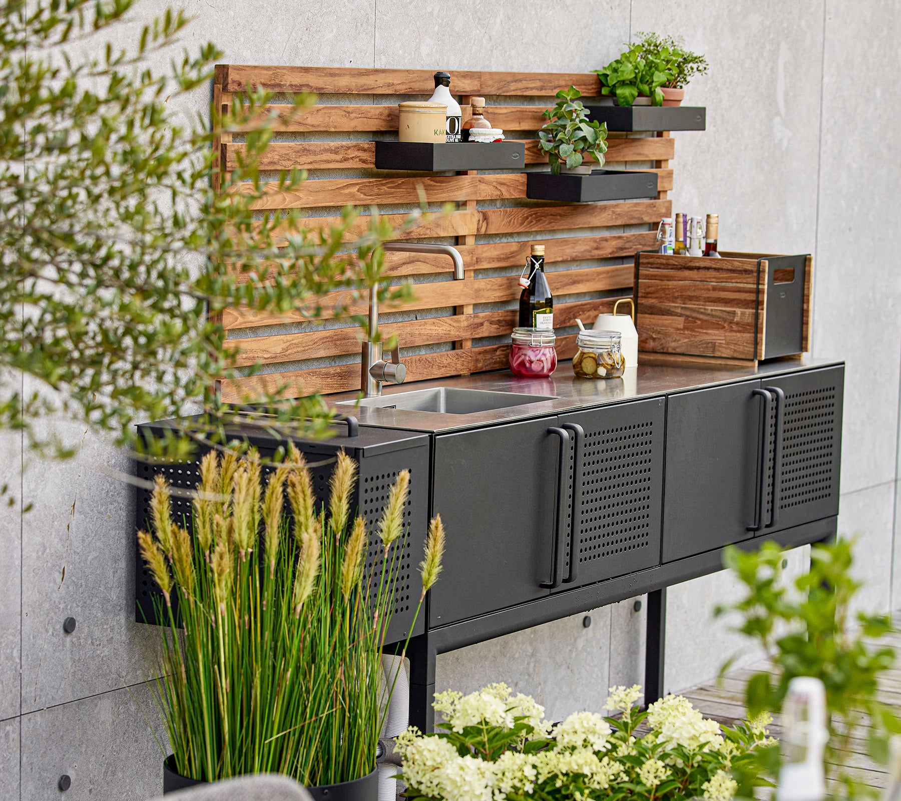 Unité de cuisine noire moderne avec un évier, des étagères en bois et diverses plantes, créant un espace de travail à la fois élégant et fonctionnel.