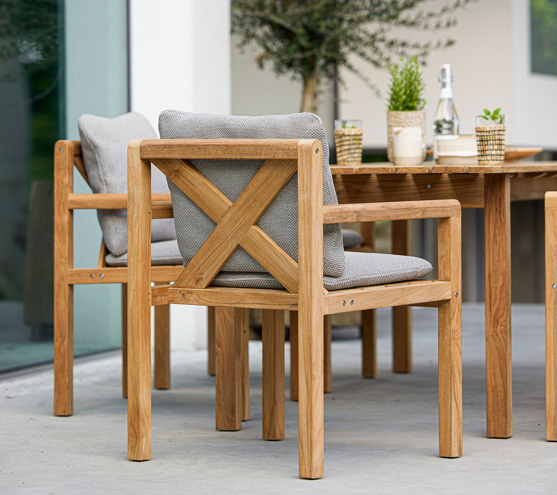 Chaises en bois avec des coussins gris et une table assortie, accompagnées de plantes décoratives et d'une bouteille, idéales pour manger en plein air.