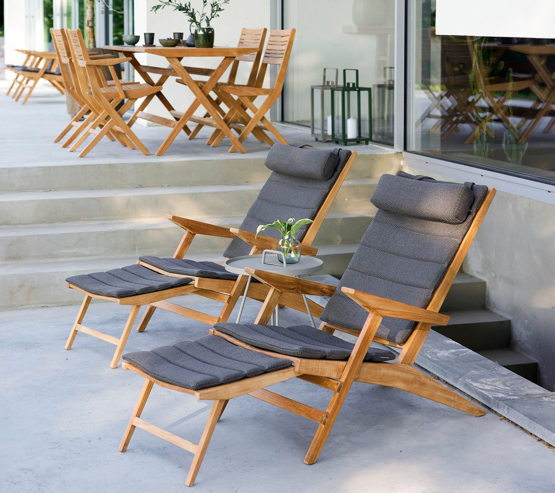 Deux chaises longues en bois avec des coussins, une petite table d'appoint et de la verdure dans un cadre extérieur élégant avec des chaises supplémentaires en arrière-plan.