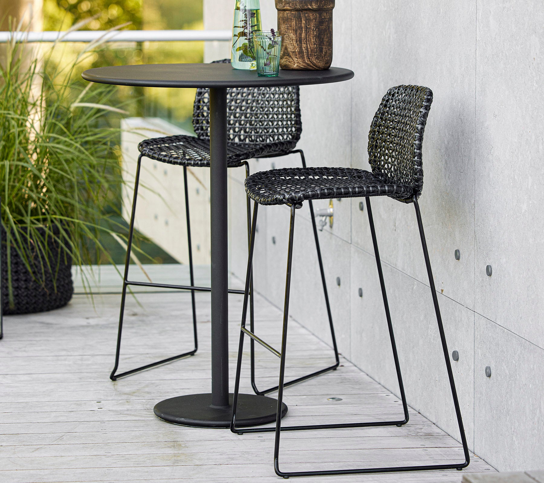 Table de bar noire élégante avec deux chaises tissées et une plante en pot, parfaite pour un repas décontracté ou pour se détendre.