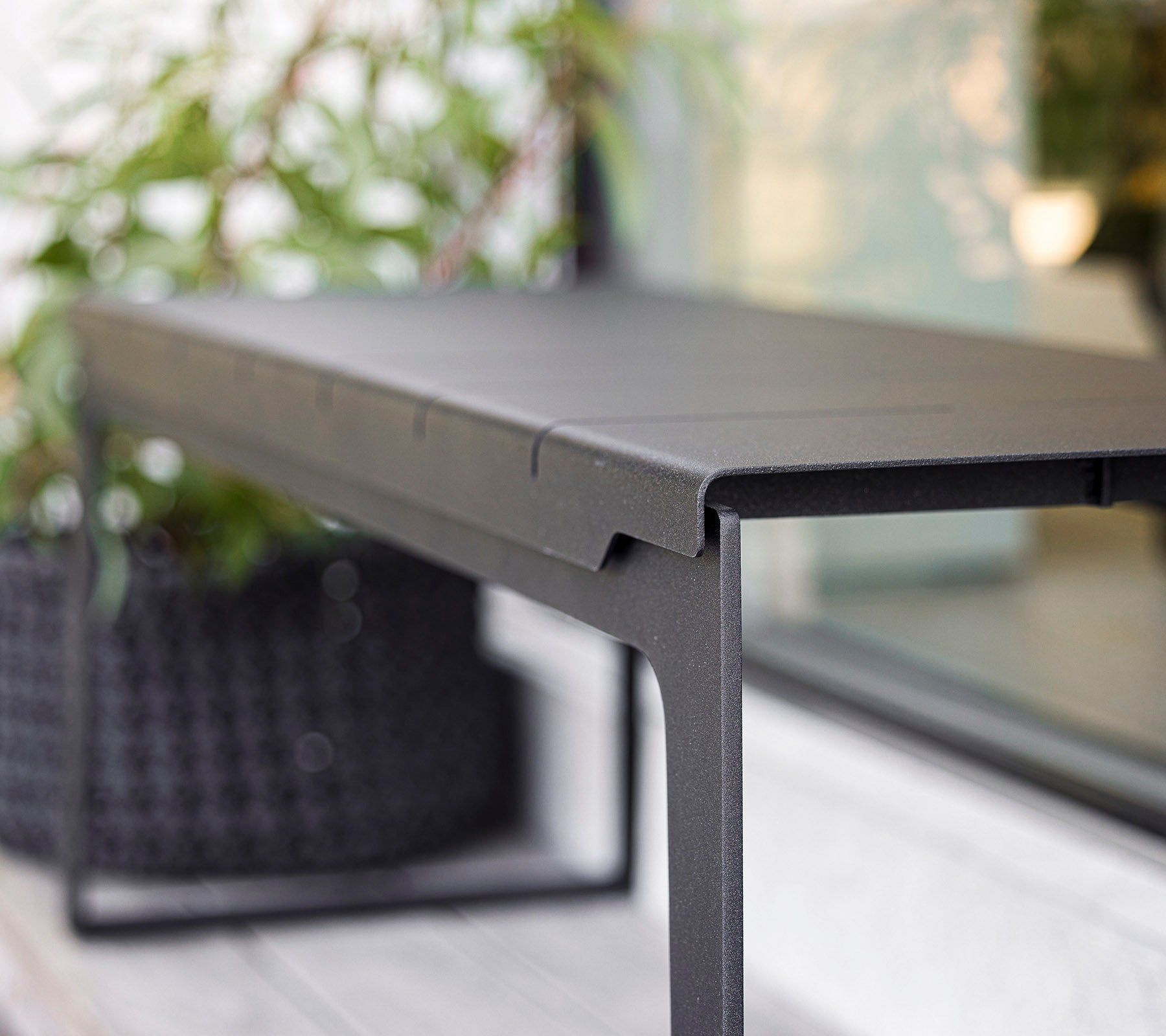 Table noire élégante au design minimaliste, présentant des lignes épurées et un cadre robuste, complété par de la verdure en arrière-plan.