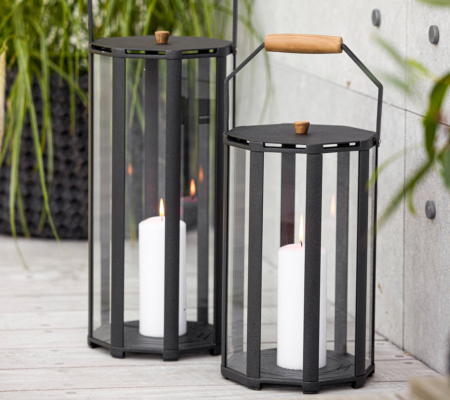Deux lanternes élégantes au design moderne, avec des panneaux en verre et une poignée en bois, contenant des bougies blanches à l'intérieur.