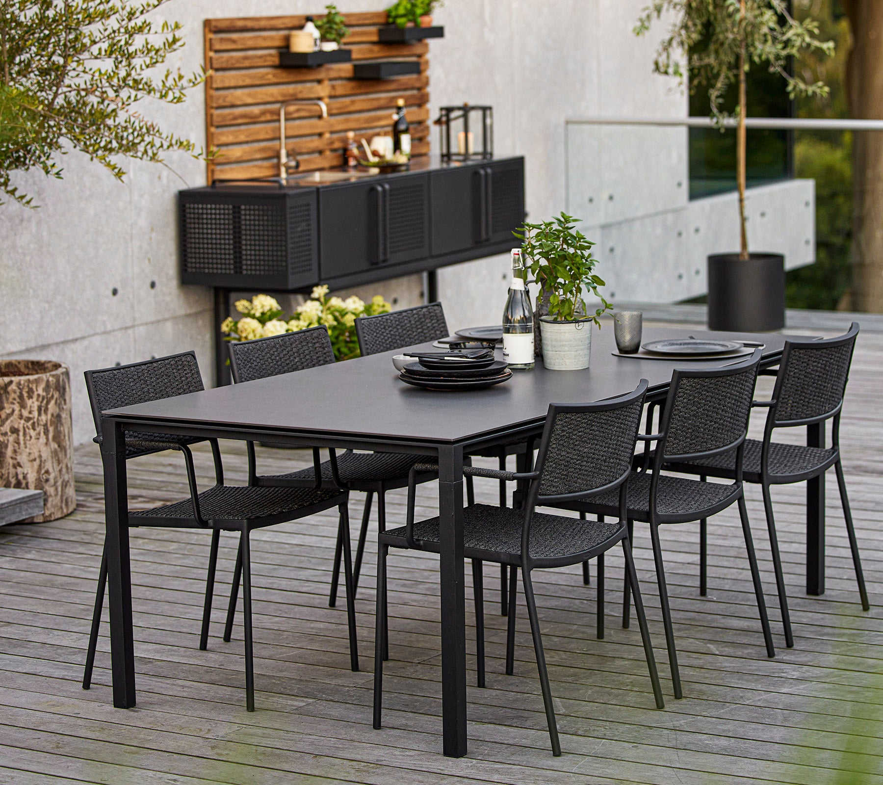 Un ensemble de salle à manger moderne comprenant une grande table rectangulaire et six chaises en osier noires, rehaussé d'éléments décoratifs et de verdure.