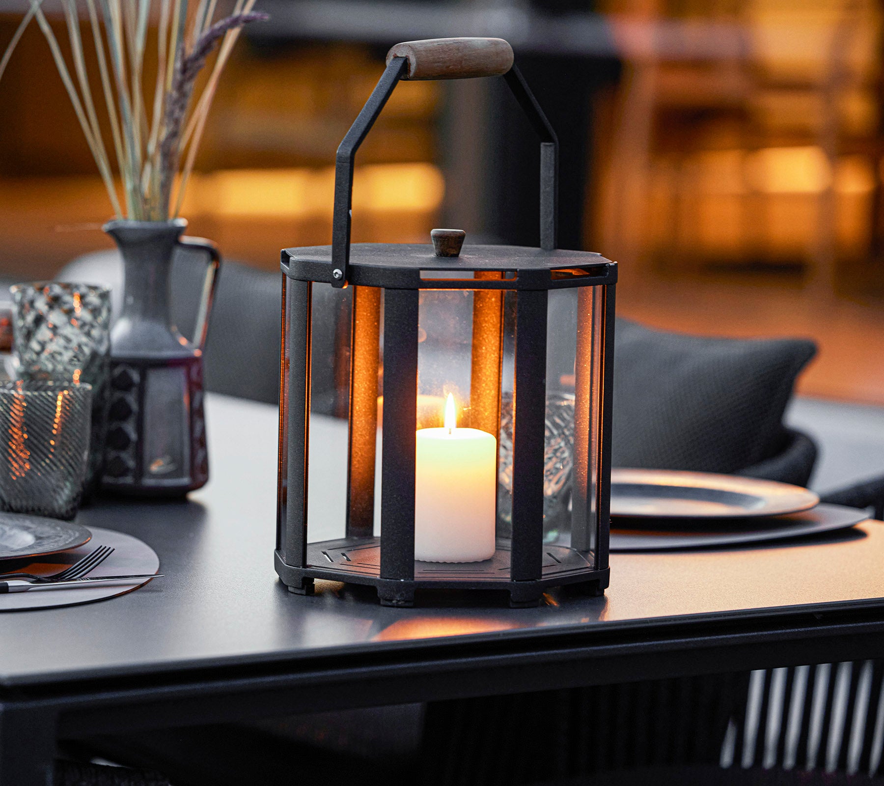 Une lanterne élégante avec une bougie à l'intérieur, dotée d'un cadre en métal et d'une poignée en bois, placée sur une table avec des éléments décoratifs autour.