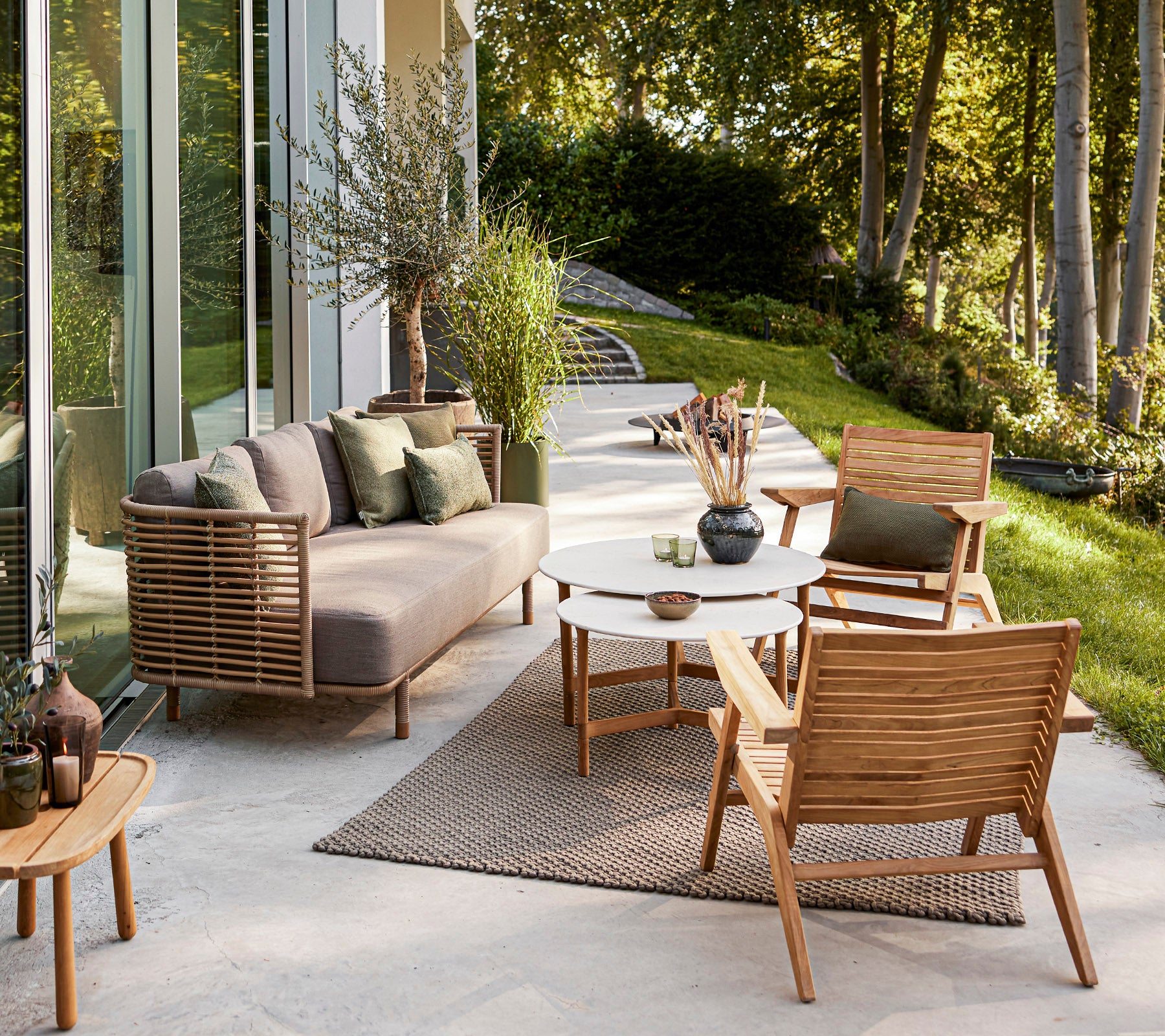 Espace extérieur élégant avec un canapé moderne, des chaises en bois, une table ronde et des plantes décoratives dans un cadre serein.