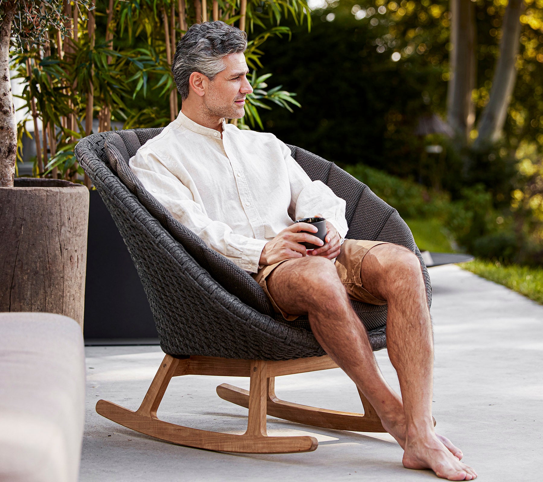 Un homme est assis confortablement dans un rocking-chair stylé, savourant une boisson dans un cadre serein, habillé décontracté dans une chemise légère et un short.