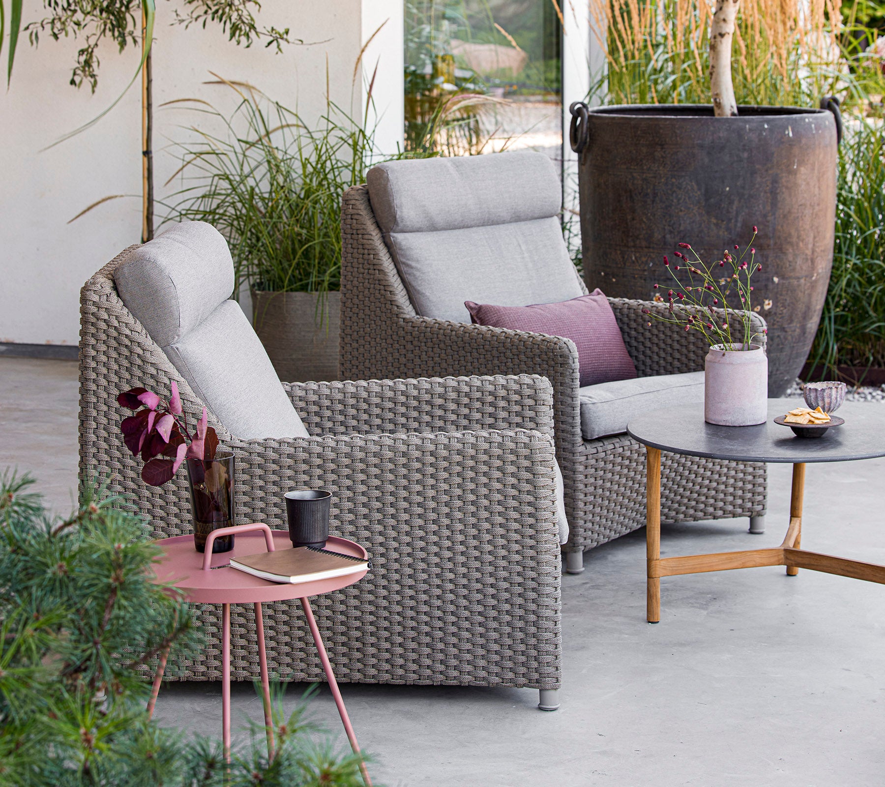 Deux chaises grises confortables avec des coussins, une table ronde avec des collations et des boissons, entourée de verdure et de décoration. Parfait pour se détendre.