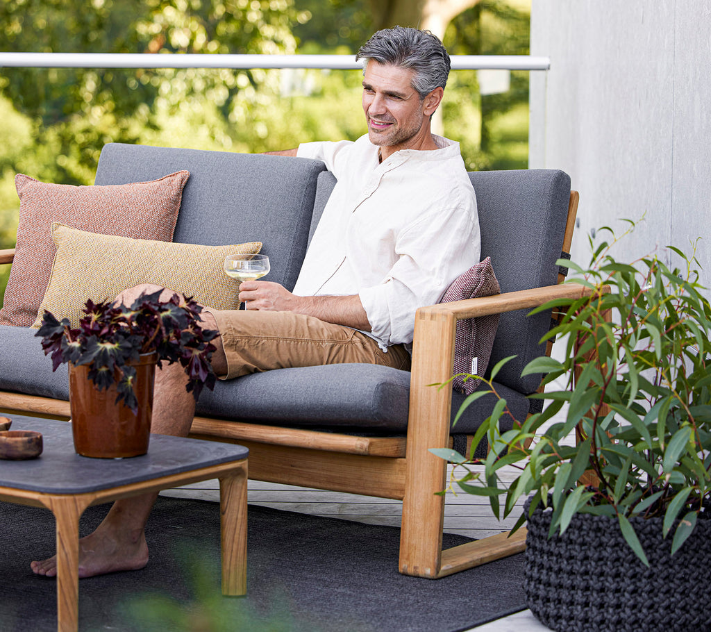 Un homme détendu est assis sur un canapé tendance avec des coussins, en train de déguster une boisson à côté d'une plante en pot et d'une table basse.