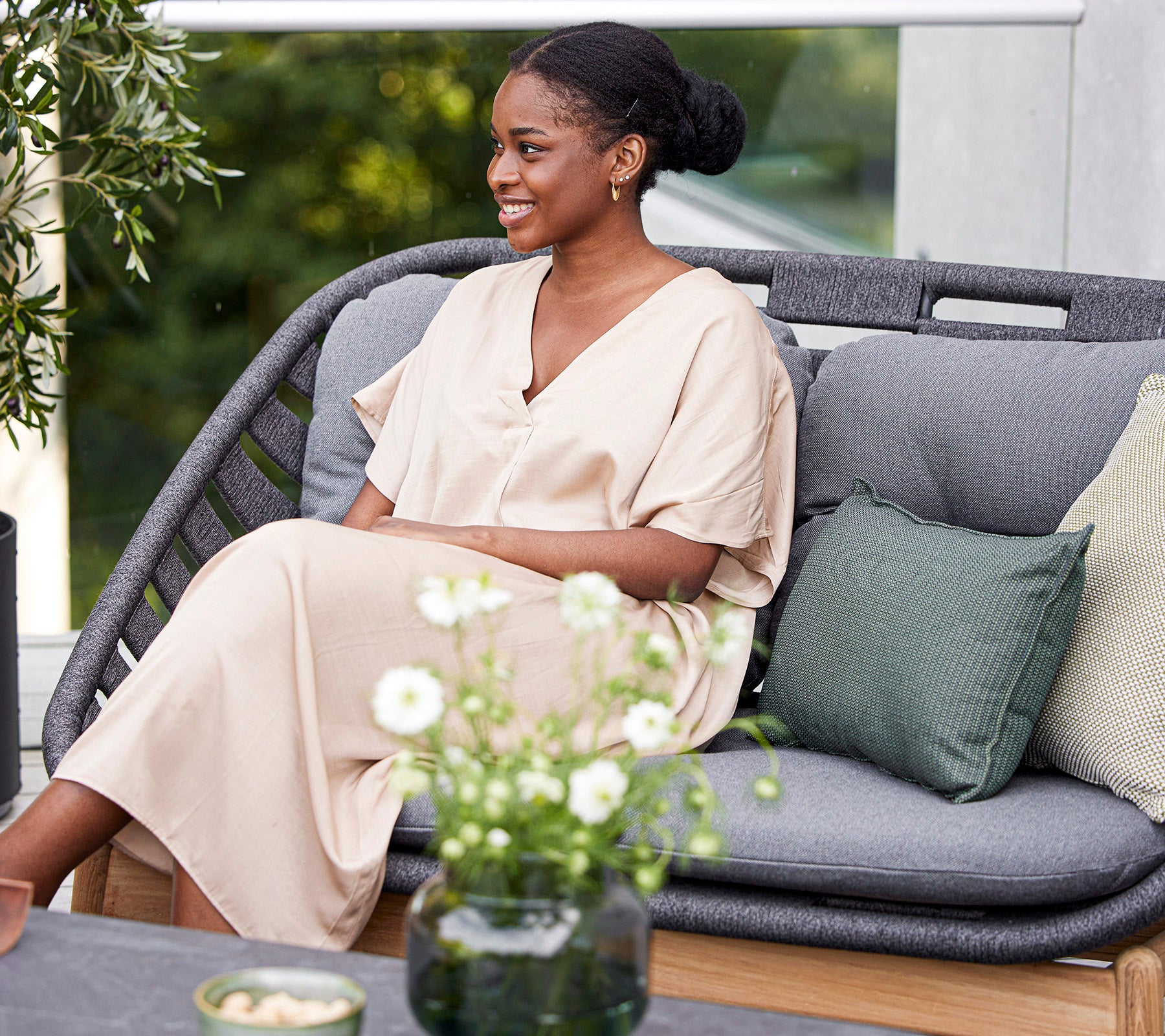 Une femme souriante en robe claire est assise sur un canapé élégant, entourée de coussins décoratifs et d'un petit arrangement floral.