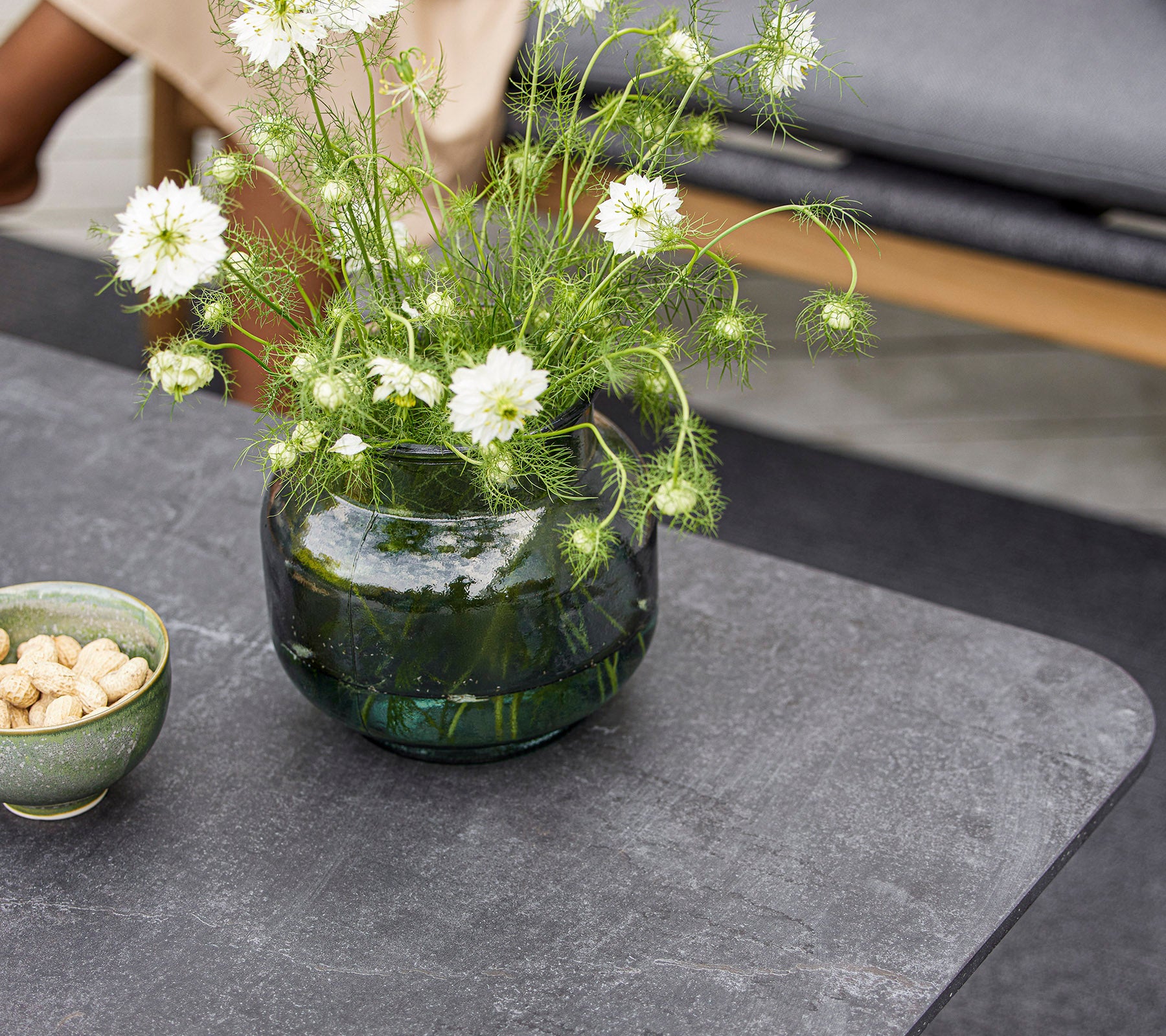 Une table moderne avec un vase en verre rond contenant des fleurs blanches et un petit bol de noix. La surface est lisse et texturée.