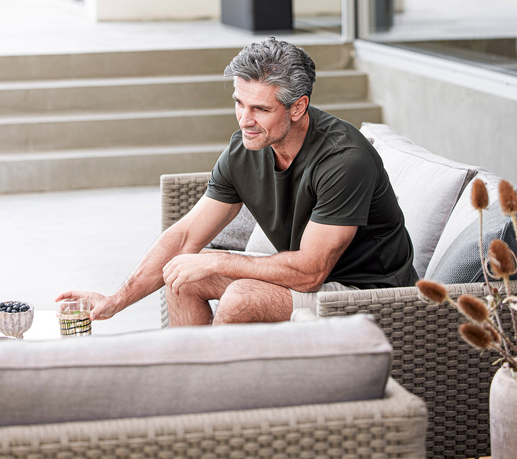 Un homme est assis confortablement sur un canapé, tenant une boisson et souriant, entouré de coussins doux et d'éléments décoratifs.