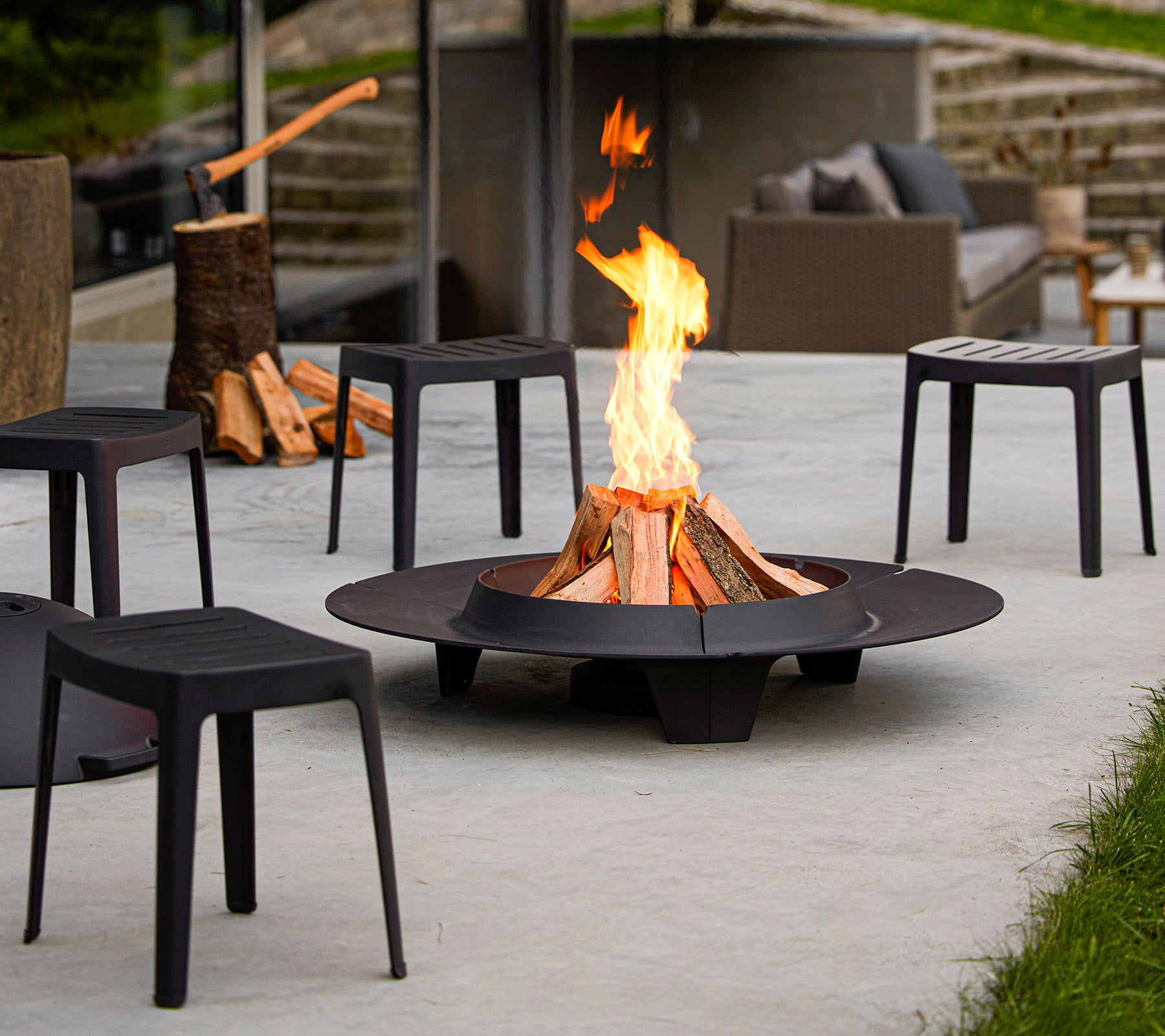 Un foyer entouré de chaises noires minimalistes, avec une flamme vive et des tas de bois de chauffage à proximité.