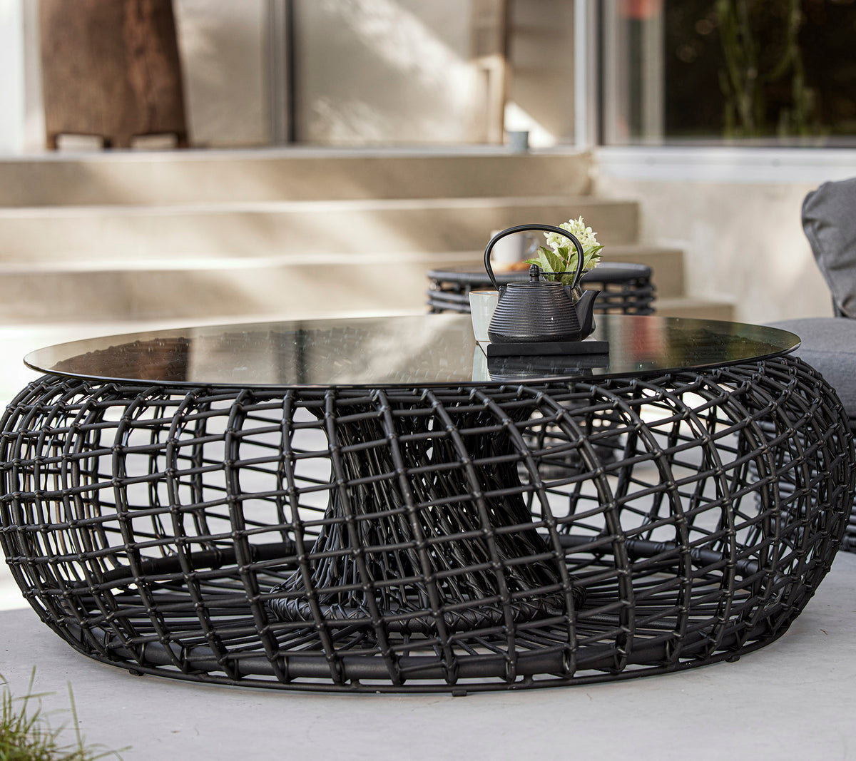 Une table basse moderne et ronde avec une base tissée en noir et un plateau en verre, comportant des objets décoratifs tels qu'une plante et un petit bol.