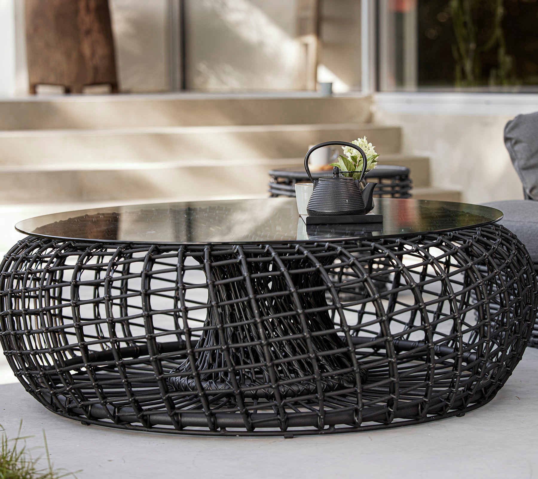 Une table basse moderne et ronde avec une base tissée en noir et un plateau en verre, comportant des objets décoratifs tels qu'une plante et un petit bol.