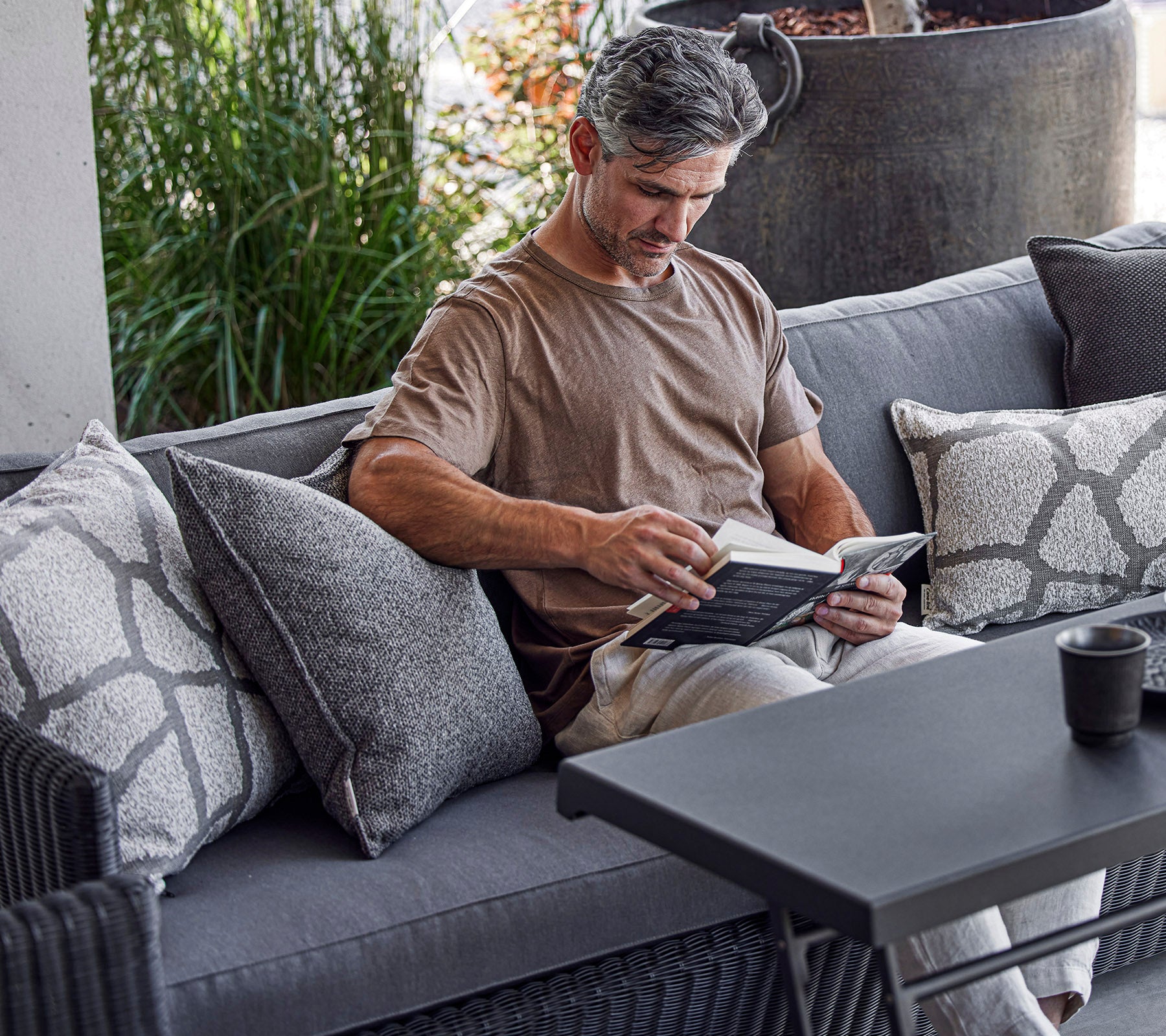 Un homme lit un livre tout en étant assis sur un canapé confortable orné de coussins décoratifs, avec de la verdure en arrière-plan.