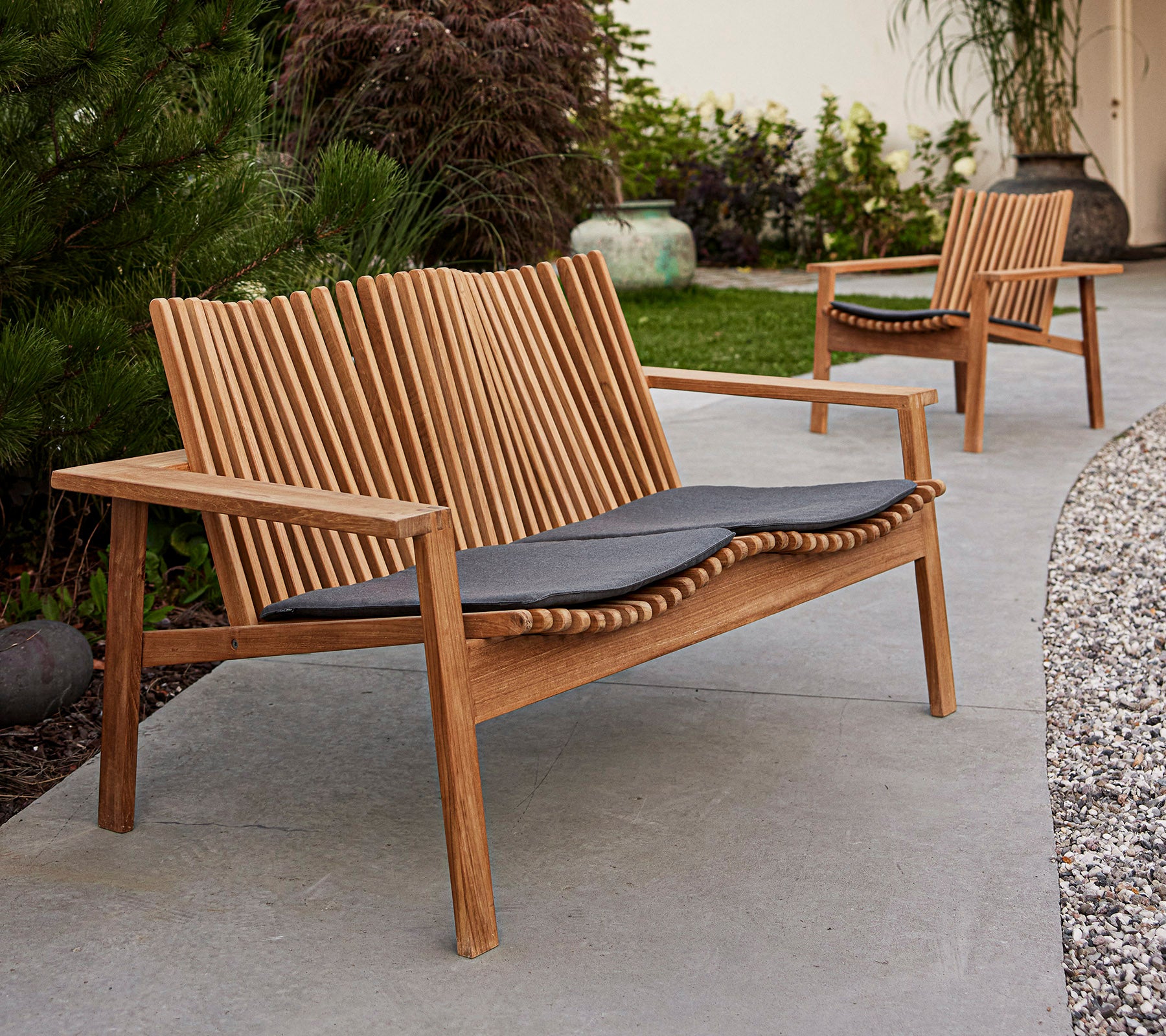 Bancs en bois modernes avec dossiers à lattes et coussins sombres, situés dans une zone paysagée avec de la verdure.