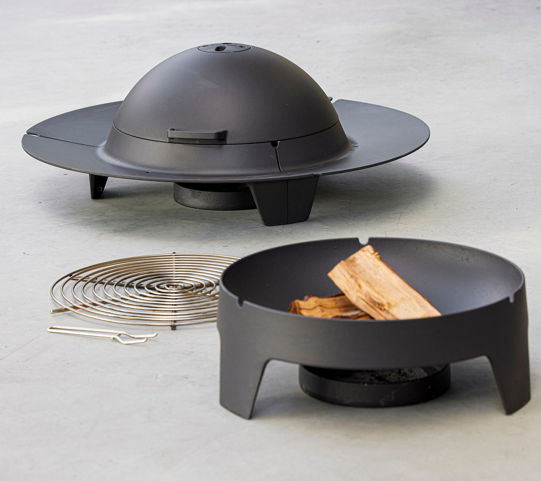 Un ensemble de foyer moderne avec un design circulaire épuré et un couvercle en dôme, accompagné d'un grille et de morceaux de bois.