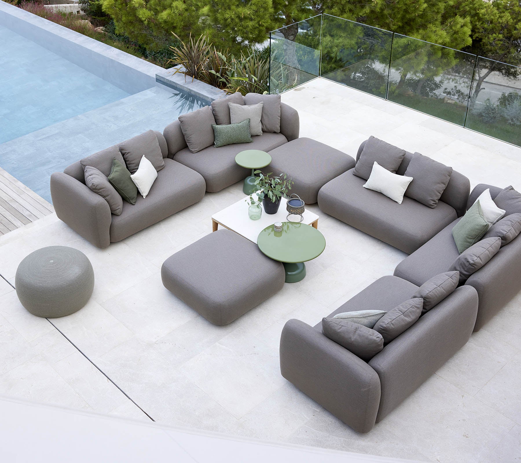 Arrangement moderne de sièges extérieurs avec des canapés gris et des tables vertes à côté d'une piscine, entouré de verdure.