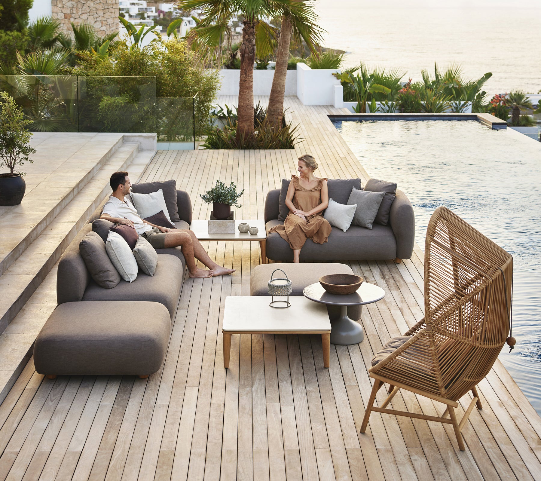 Espace lounge moderne en extérieur avec des canapés gris, une table blanche et une chaise en rotin.