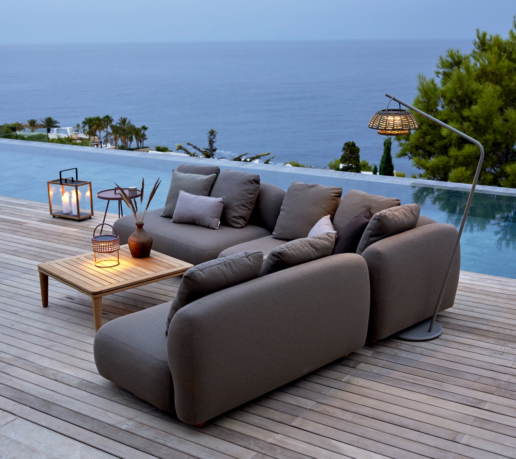 Sofa gris avec table en bois et lanternes au bord de la piscine surplombant l'océan.
