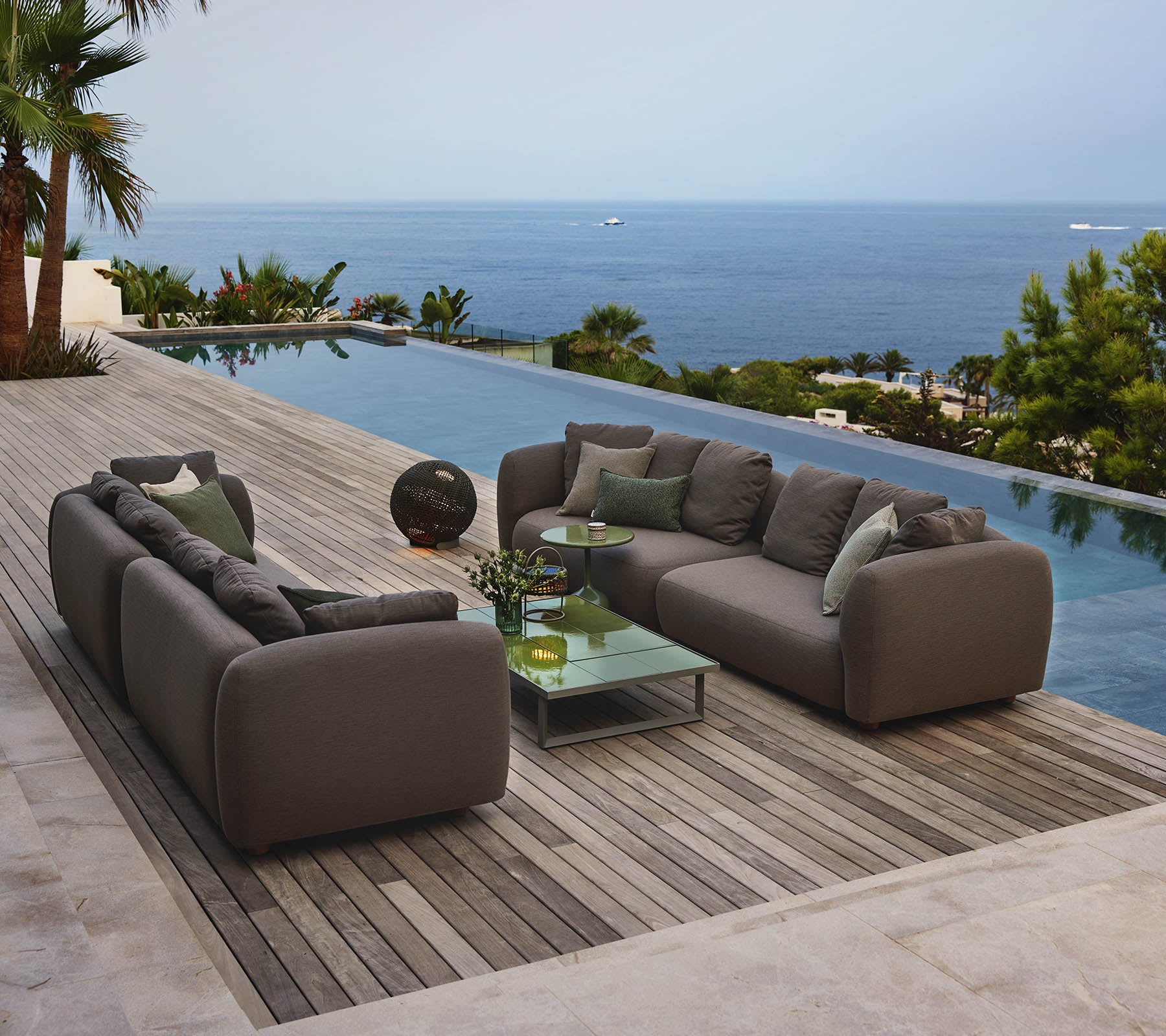Mobilier de salon moderne disposé autour d'une table basse en verre, surplombant une piscine sereine et une vaste vue sur l'océan, entouré de verdure.