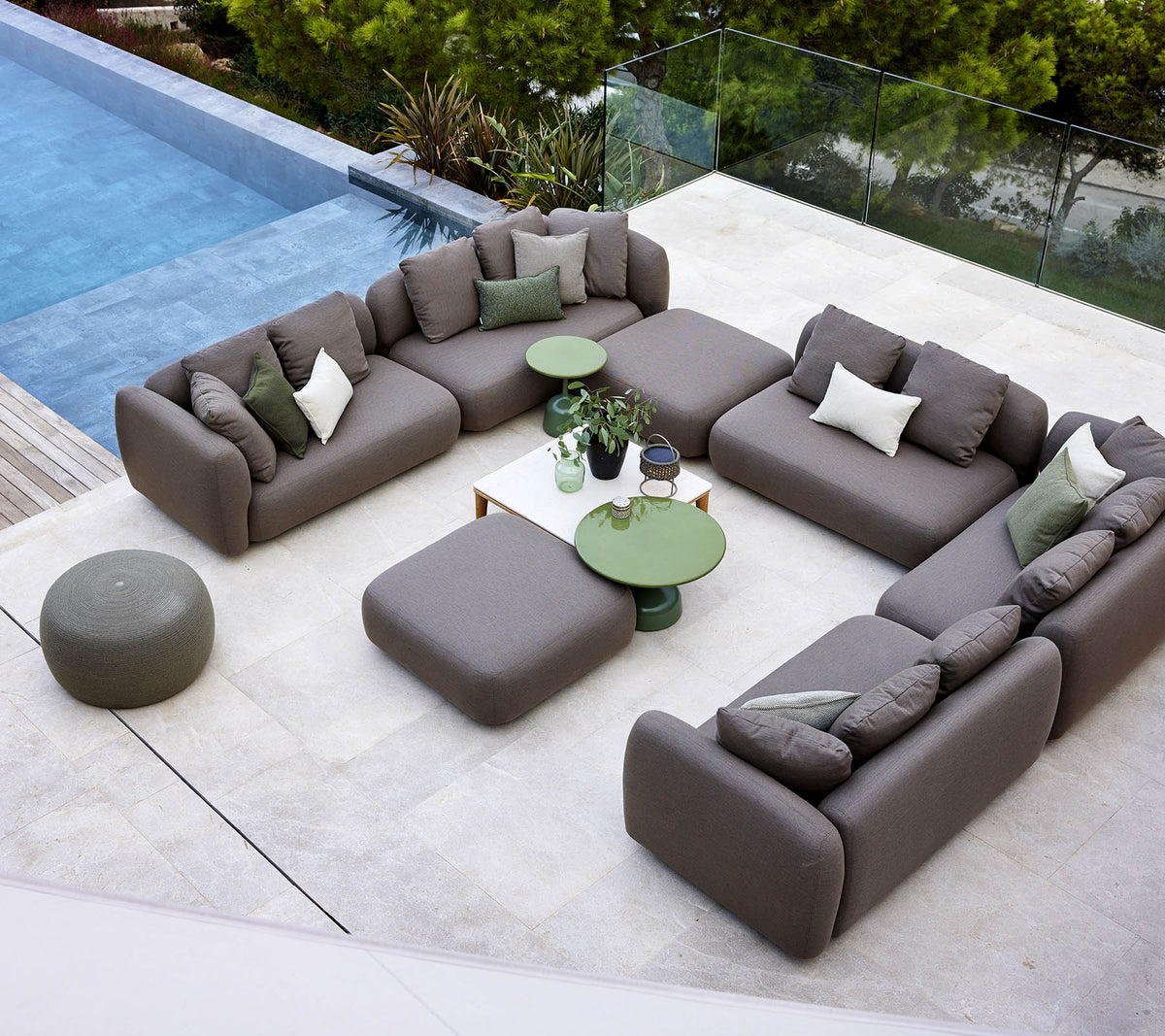Un aménagement moderne de salon avec un canapé sectionnel, un pouf rond et des tables d'appoint vertes, entouré de verdure et d'une piscine paisible.