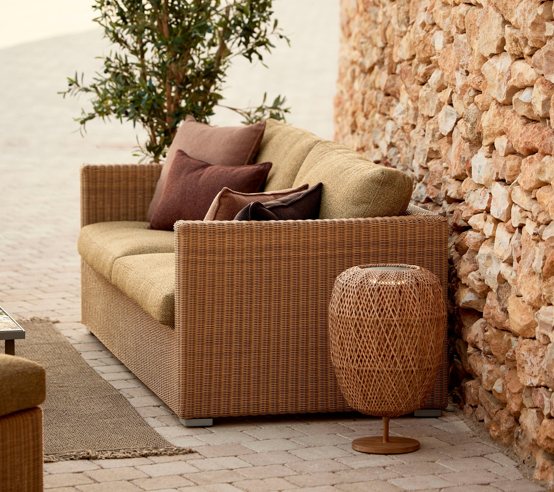 Un canapé en osier confortable avec des coussins, complété par une table d'appoint tissée élégante, situé contre un mur en pierre et de la verdure luxuriante.