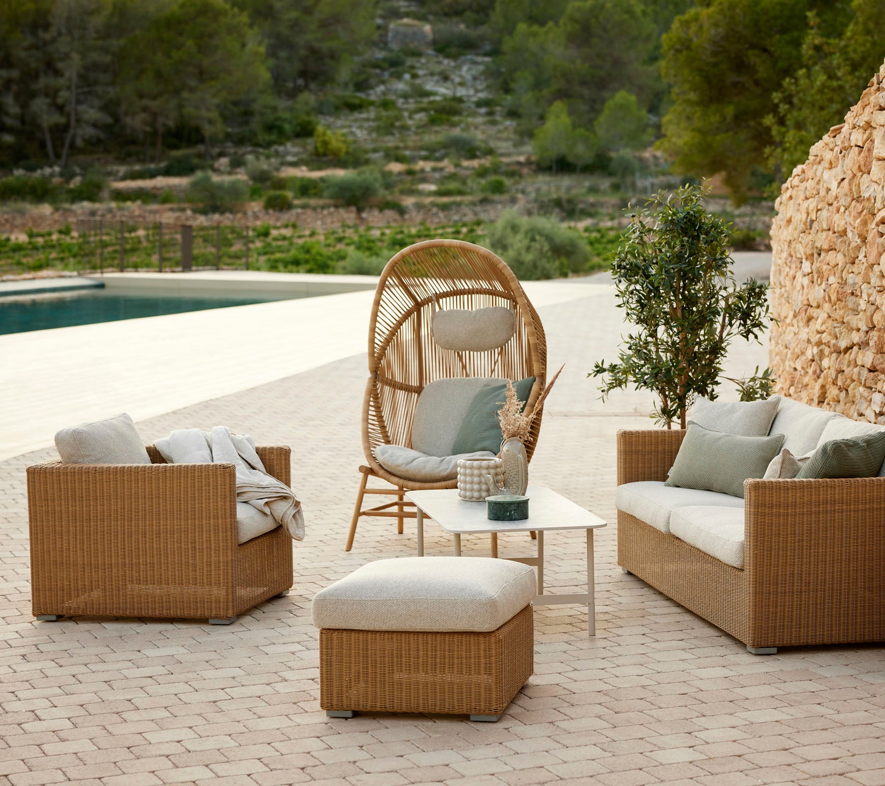 Arrangement de sièges d'extérieur confortable avec des fauteuils en rotin, un fauteuil suspendu circulaire, un pouf et une petite table parmi la verdure.